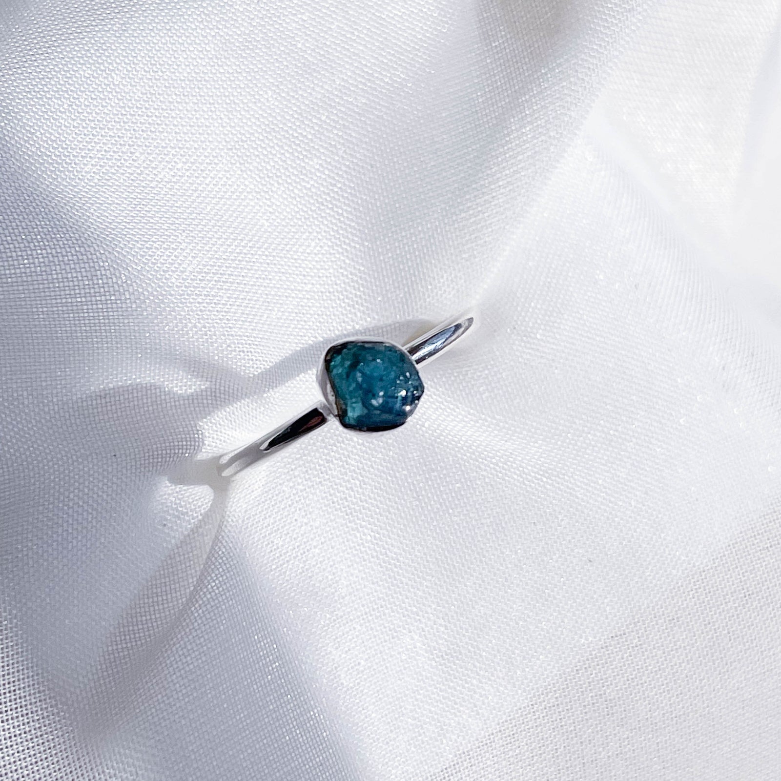 The Healing Hedge Witch's Blue Tourmaline Stackable Rough Crystal Ring, featuring an oval blue gemstone, is displayed on softly textured white fabric under natural light.