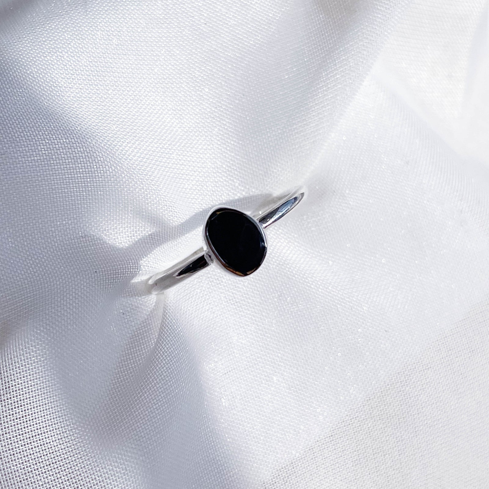 The Healing Hedge Witch’s Shungite Stackable Rough Crystal Ring features a small, oval Shungite stone—known for its protective properties—set in silver and displayed on white textured fabric.