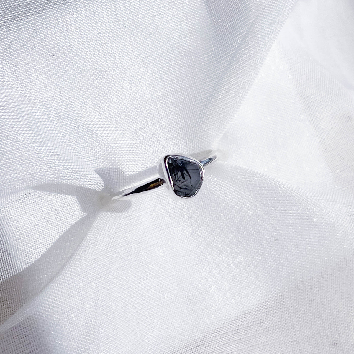 The Healing Hedge Witch Tourmalinated Quartz Stackable Rough Crystal Ring features a minimalist silver band with an irregular dark stone, showcased on textured white fabric.
