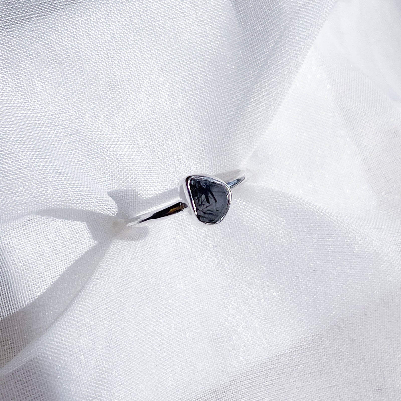 The Healing Hedge Witch Tourmalinated Quartz Stackable Rough Crystal Ring features a minimalist silver band with an irregular dark stone, showcased on textured white fabric.