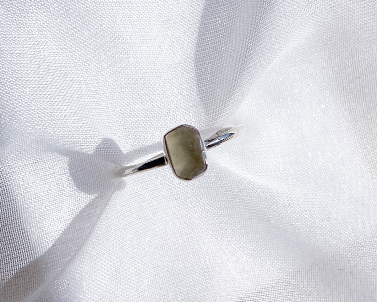 The Healing Hedge Witch’s Libyan Desert Glass Stackable Rough Crystal Ring, featuring a faceted hexagonal pale green stone, rests on softly crumpled white fabric, illuminated by bright light.