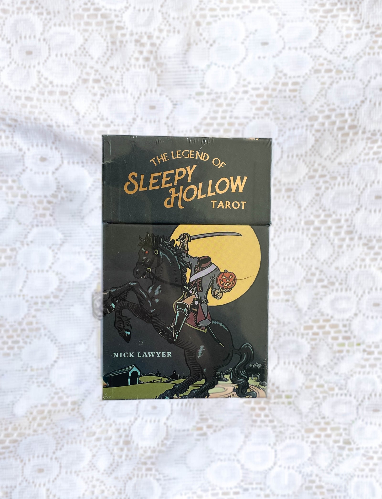 A tarot deck box, The Legend of Sleepy Hollow Tarot by The Healing Hedge Witch, displays the Headless Horseman with a pumpkin against a yellow moon, set on a white lace surface.