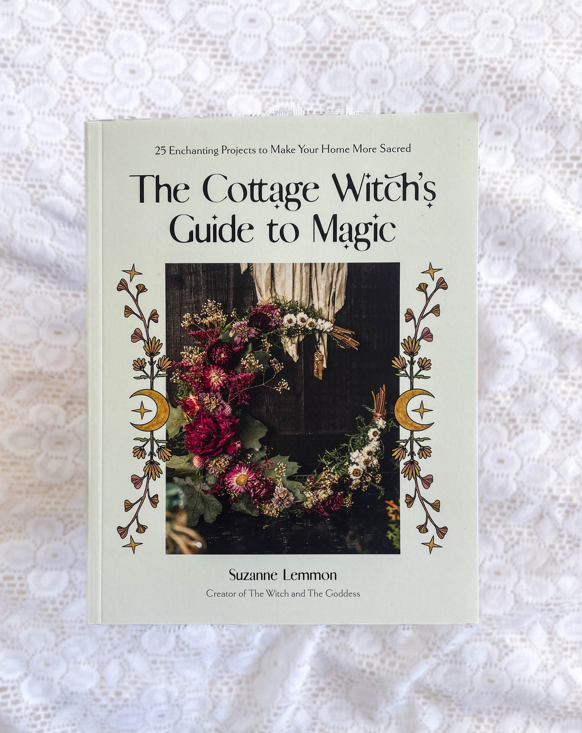 The Healing Hedge Witch’s Cottage Witch's Guide To Magic: 25 Enchanting Home Projects rests on white lace. The cover displays a floral wreath, crescent moons, and a subtitle about enchanting projects for a sacred home.