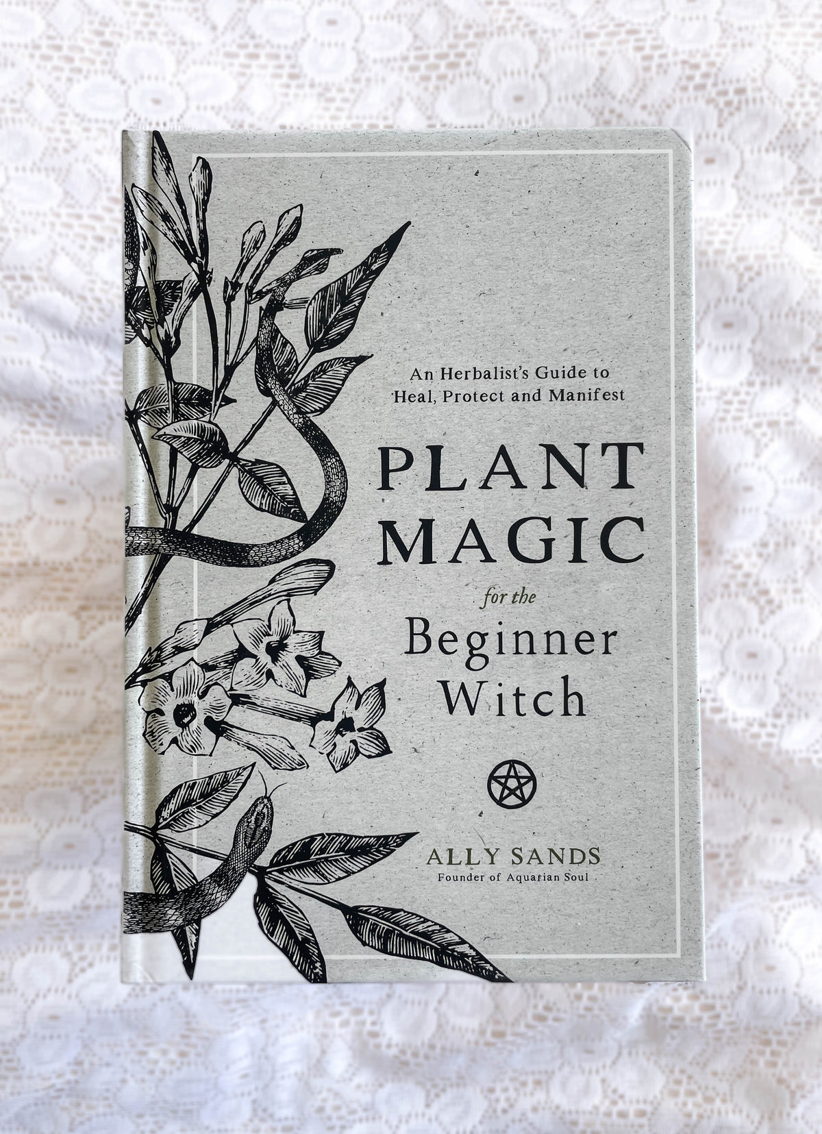 Plant Magic For Beginner Witches: Guide To Heal & Protect by The Healing Hedge Witch is displayed on white lace, featuring a cover with botanical illustrations and a subtitle focused on healing and protection.