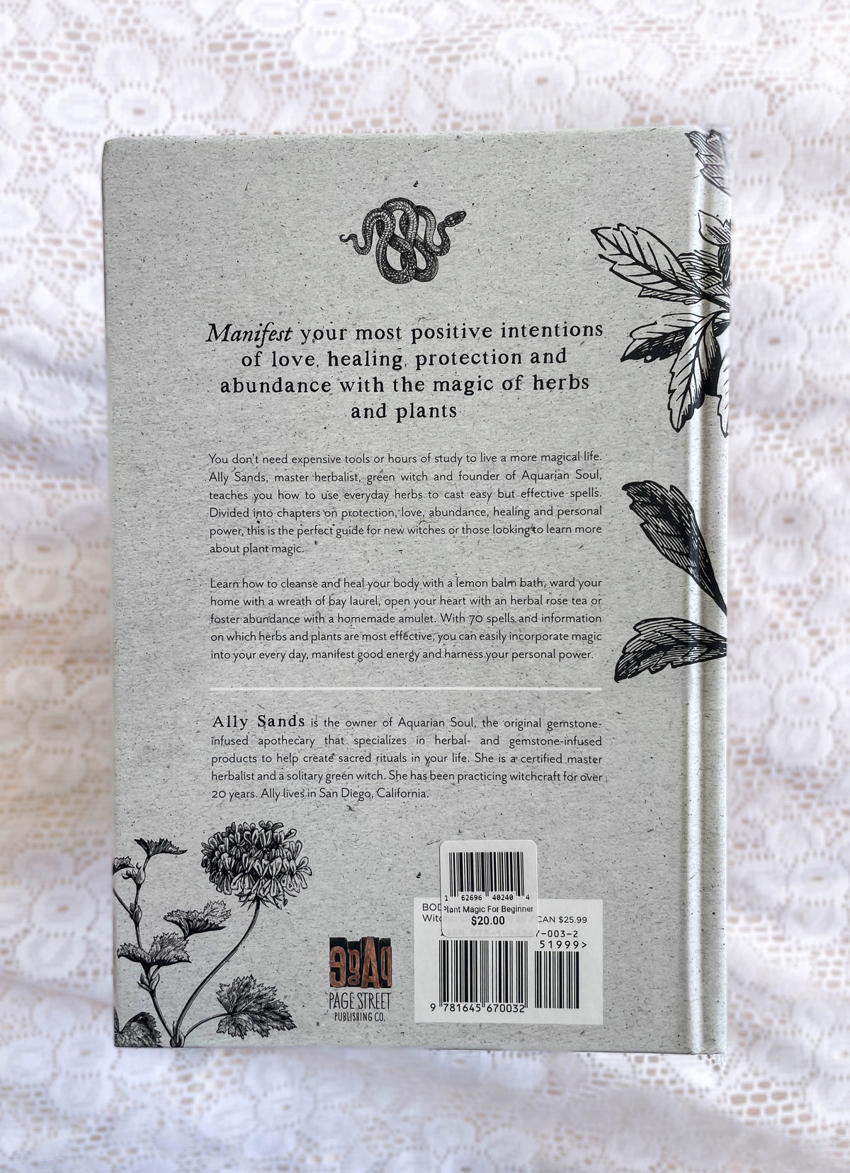 The back cover of *Plant Magic For Beginner Witches: Guide To Heal & Protect* by The Healing Hedge Witch rests on white lace, with botanical and snake designs, barcode, price, and text on manifesting intentions with herbs and plants.