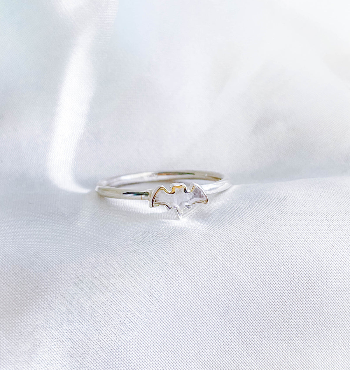 The Healing Hedge Witch's Rainbow Moonstone Bat Crystal Stackable Ring, featuring a bat-shaped charm, is displayed on soft white fabric.