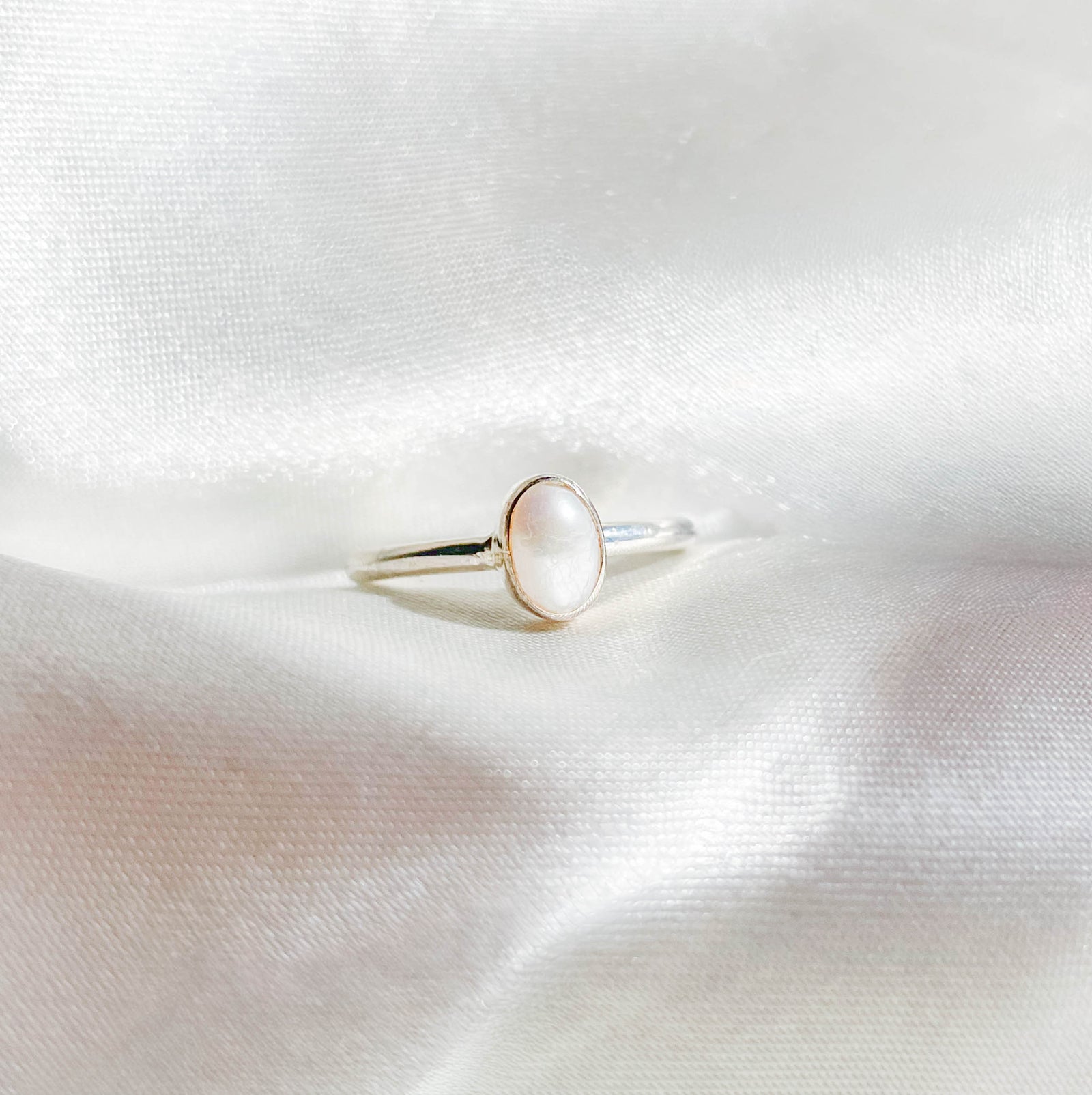 The Healing Hedge Witch Mother of Pearl Stackable Ring features a delicate silver band with an oval mother of pearl gemstone, elegantly displayed on soft white satin.