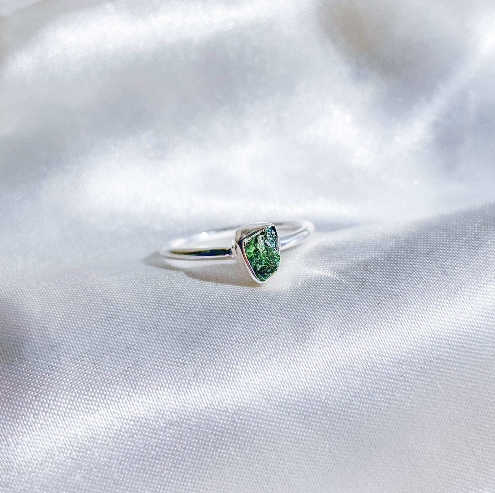 The Healing Hedge Witch’s Green Tourmaline Stackable Rough Crystal Ring is shown on white silky fabric, its green gemstone softly reflecting light.