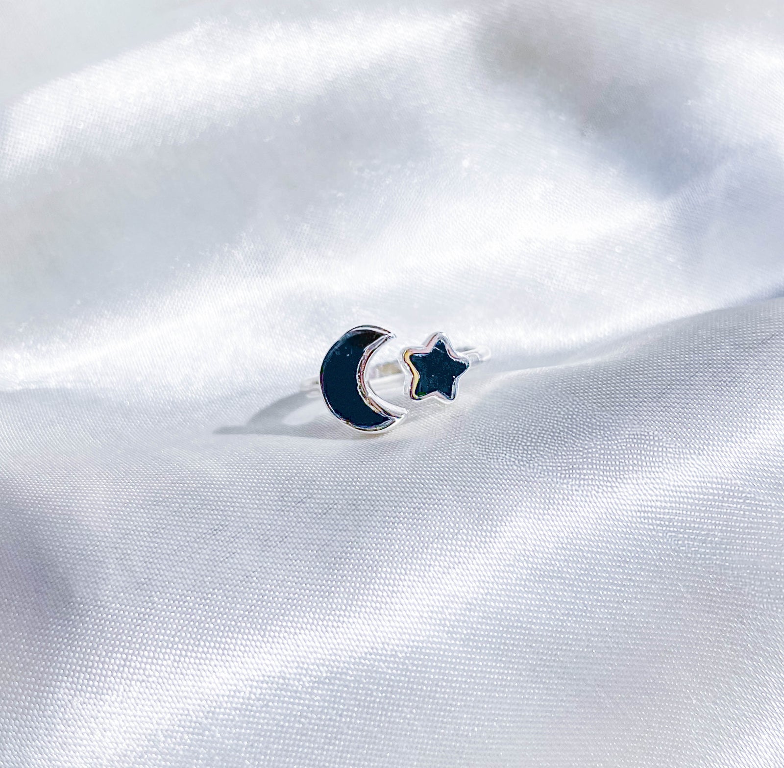 The Healing Hedge Witch’s Black Tourmaline Moon & Star Stackable Crystal Ring rests on smooth, white satin fabric.