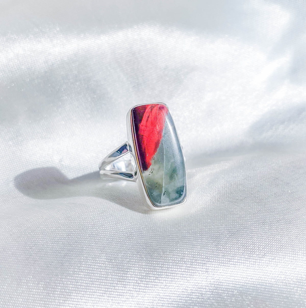 The Bloodstone Adjustable Ring by The Healing Hedge Witch features a rectangular stone in red, green, and gray hues set in silver, shown on a shiny white fabric background.