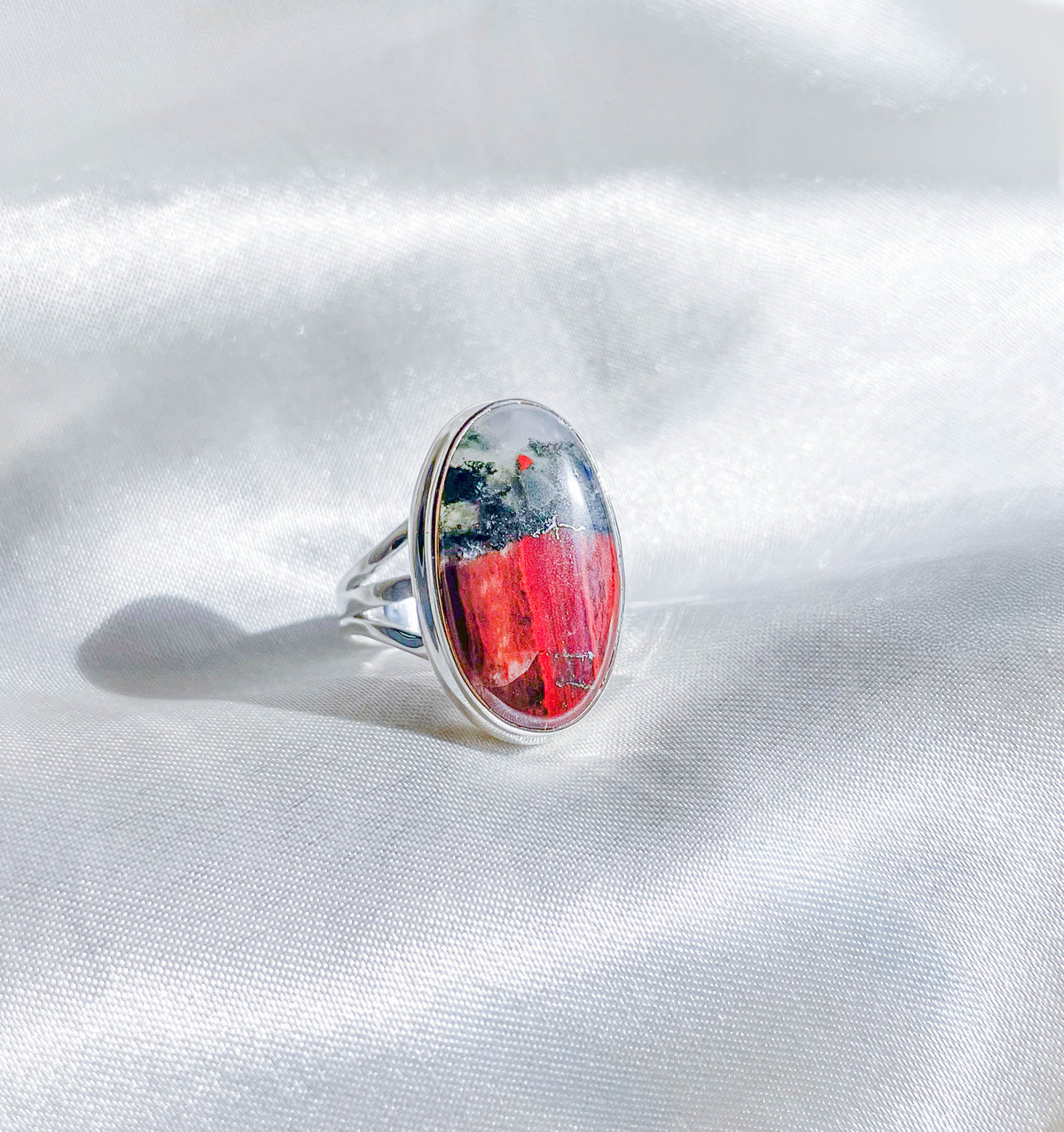 The Bloodstone Adjustable Ring by The Healing Hedge Witch features an oval silver band and a polished gemstone with red, black, and white patterns, shown on a shiny white fabric background.
