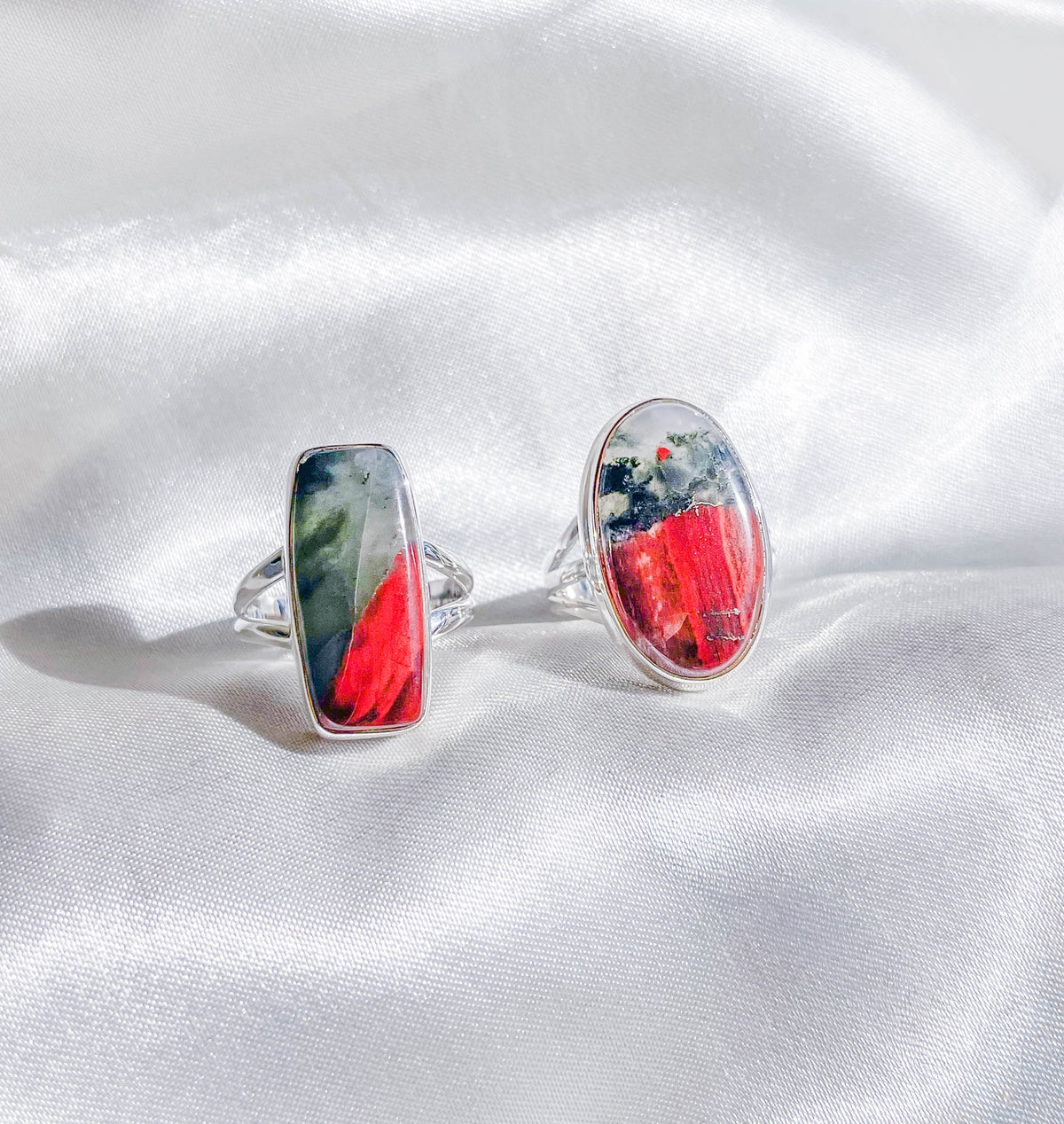 Two Bloodstone Adjustable Rings by The Healing Hedge Witch, featuring abstract red, black, and white stone settings, are displayed on white silky fabric with bright natural lighting enhancing their colors and details.