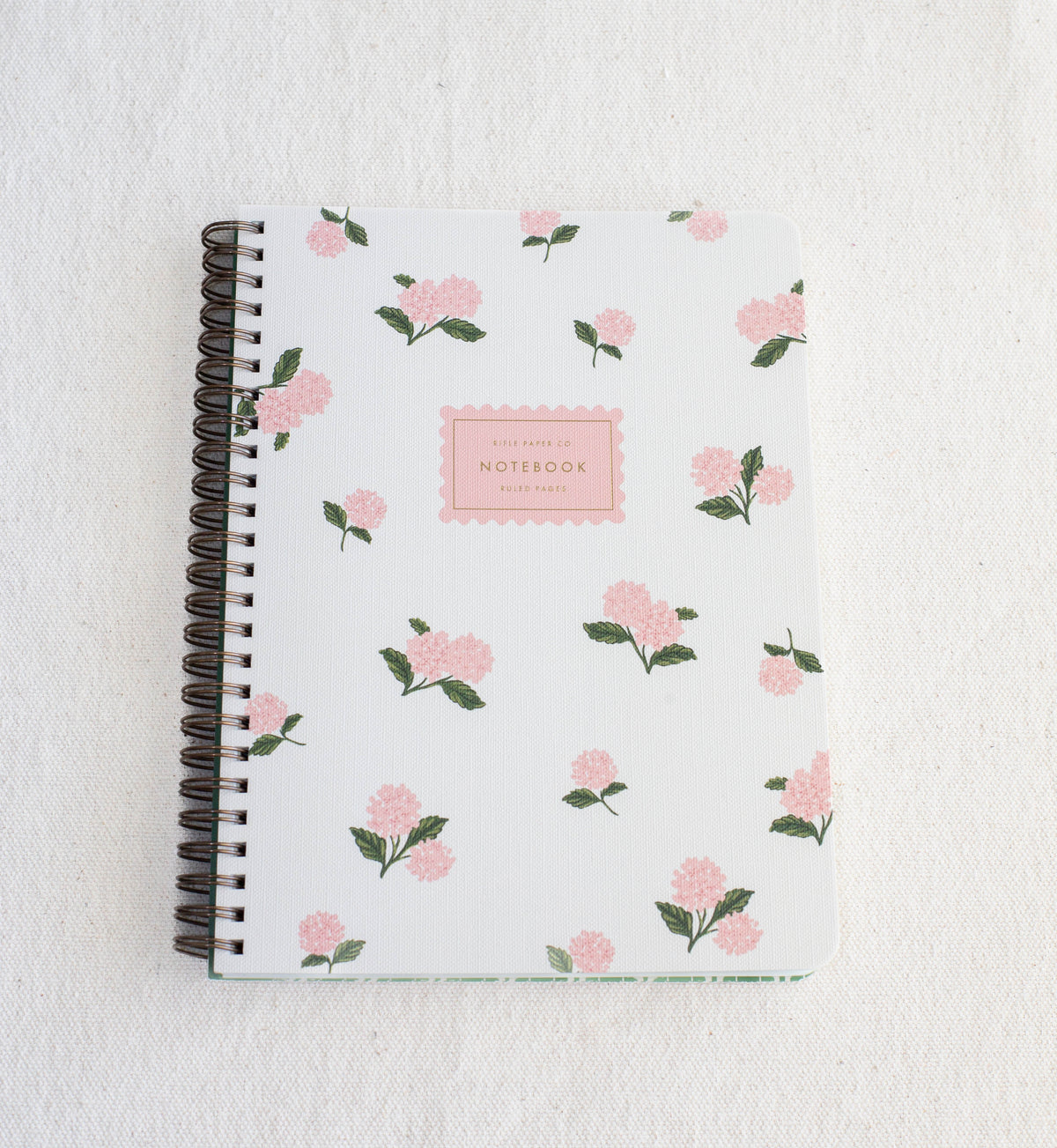 The Healing Hedge Witch's Pink Hydrangea Spiral Notebook features a white cover with pink flowers and green leaves, spiral binding, and a center label reading "NOTEBOOK" in pink, displayed on a textured white surface.