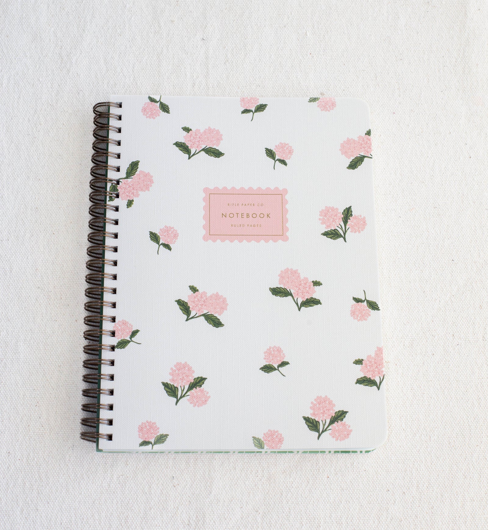 The Healing Hedge Witch's Pink Hydrangea Spiral Notebook features a white cover with pink flowers and green leaves, spiral binding, and a center label reading "NOTEBOOK" in pink, displayed on a textured white surface.