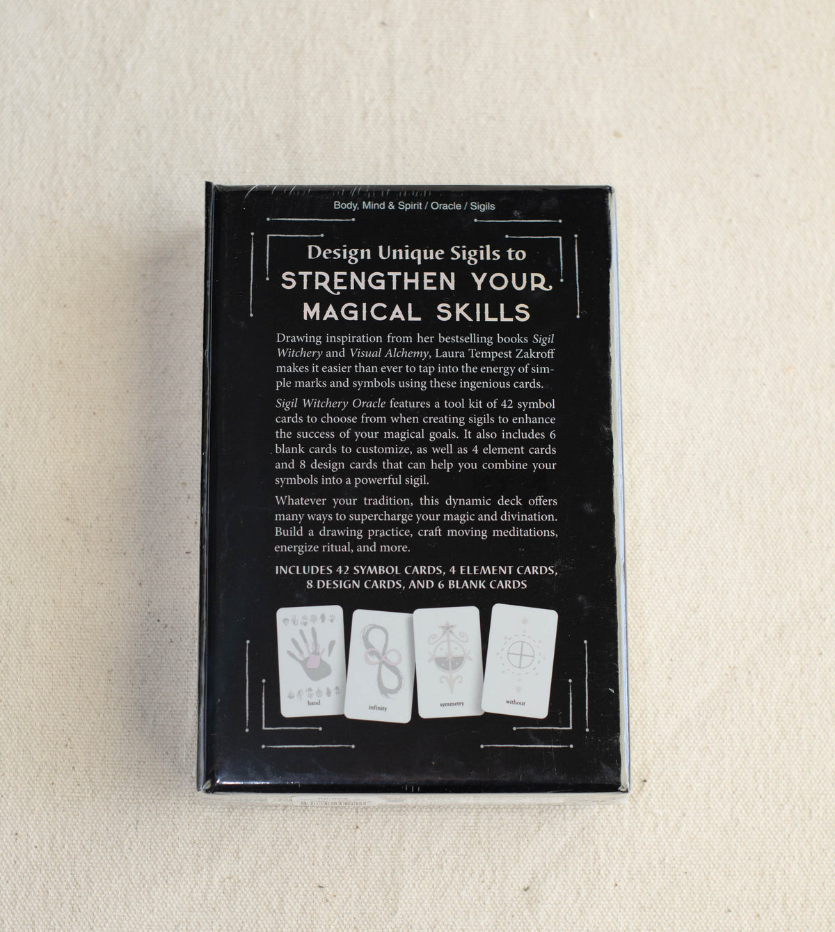 A black box with white text titled Sigil Witchery Oracle by The Healing Hedge Witch. The box lists contents and shows cards with distinct symbols on a light fabric background.