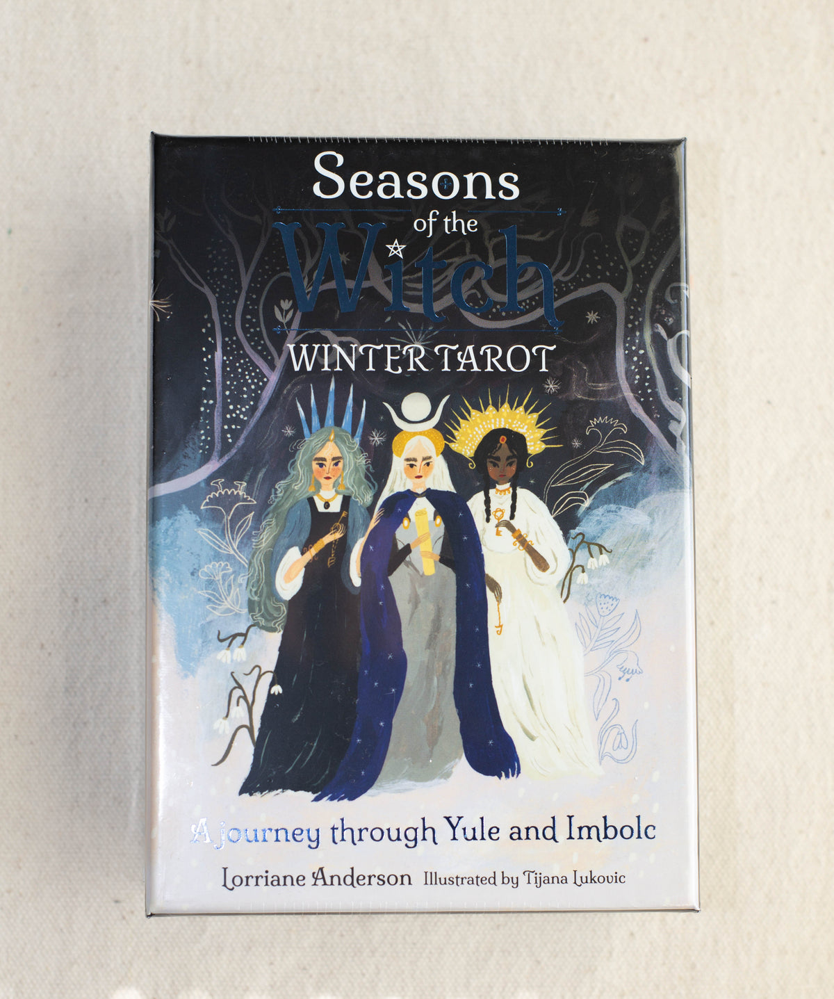 Box cover of The Healing Hedge Witch’s Seasons of the Witch Winter Tarot, featuring three women in dark, blue, and white robes under moonlight, holding candles amid winter trees.