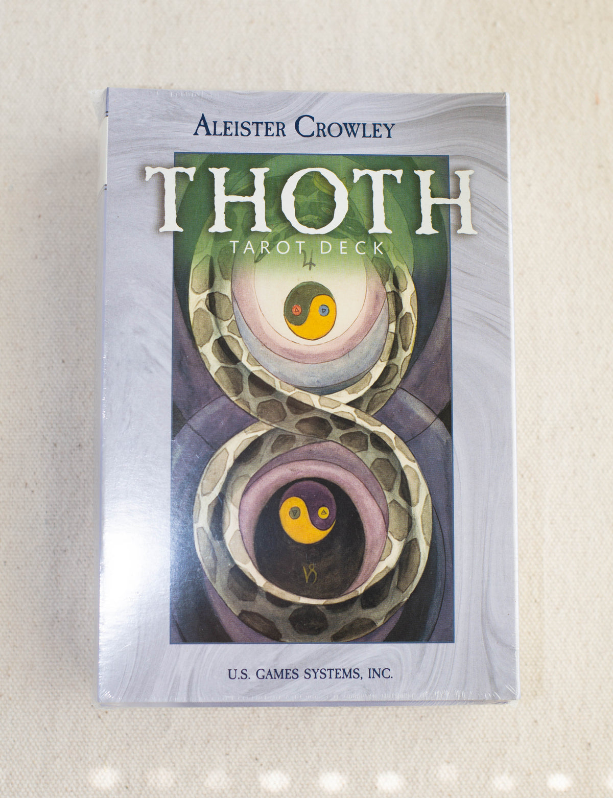 A box of the Crowley Thoth Tarot Deck — Premier Edition by The Healing Hedge Witch, with abstract art of two entwined snakes and yin-yang symbols on the cover, displayed on a light fabric background.