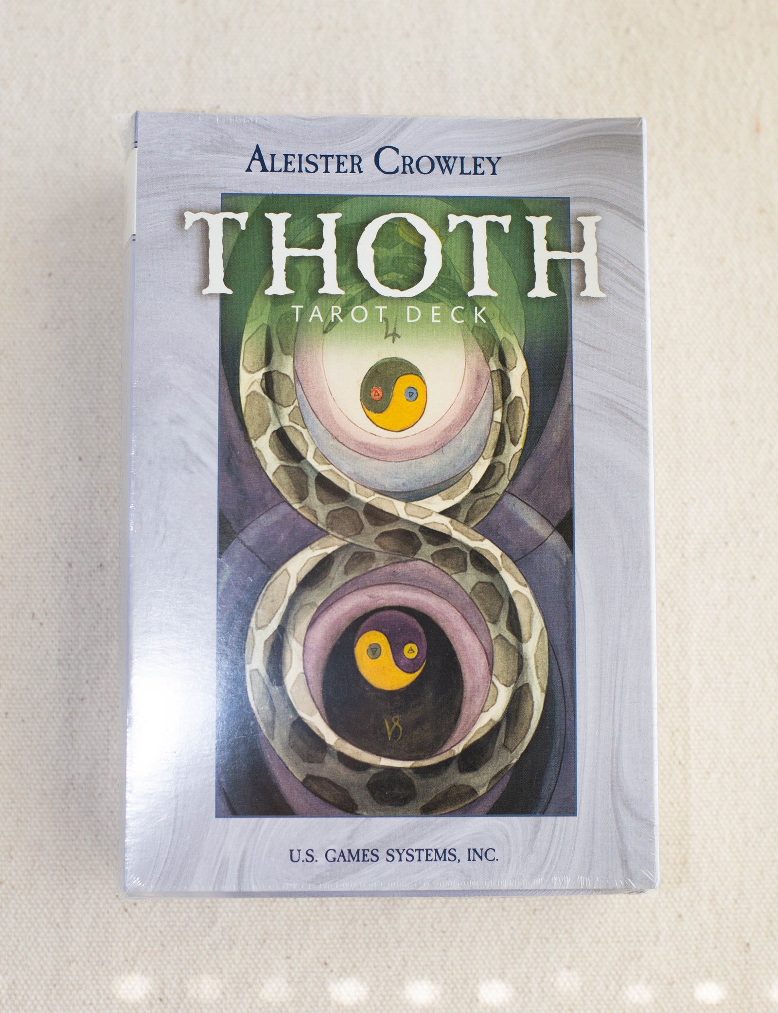 A box of the Crowley Thoth Tarot Deck — Premier Edition by The Healing Hedge Witch, with abstract art of two entwined snakes and yin-yang symbols on the cover, displayed on a light fabric background.