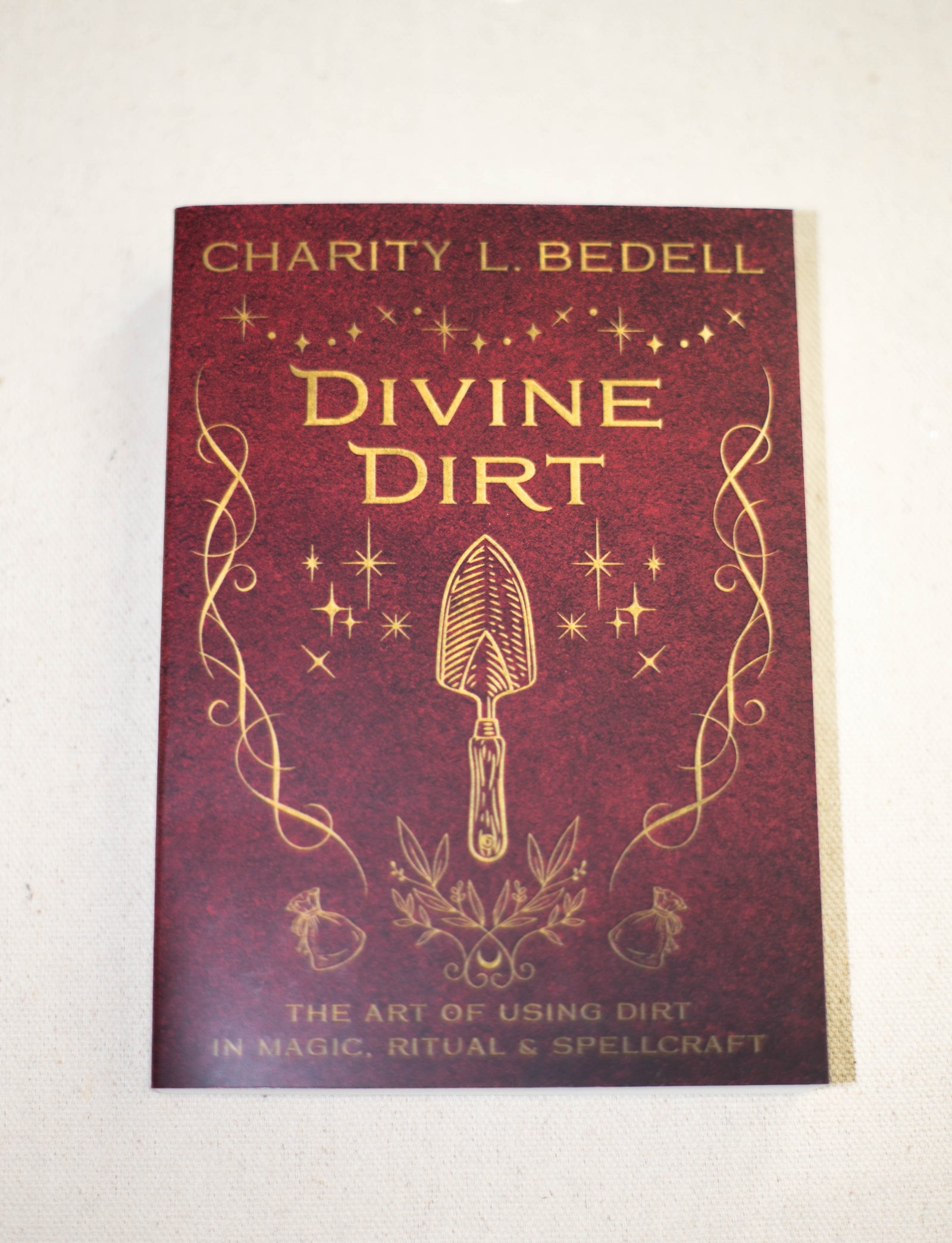 Divine Dirt by The Healing Hedge Witch is a book with a maroon cover adorned with gold mystical designs and a central trowel illustration.