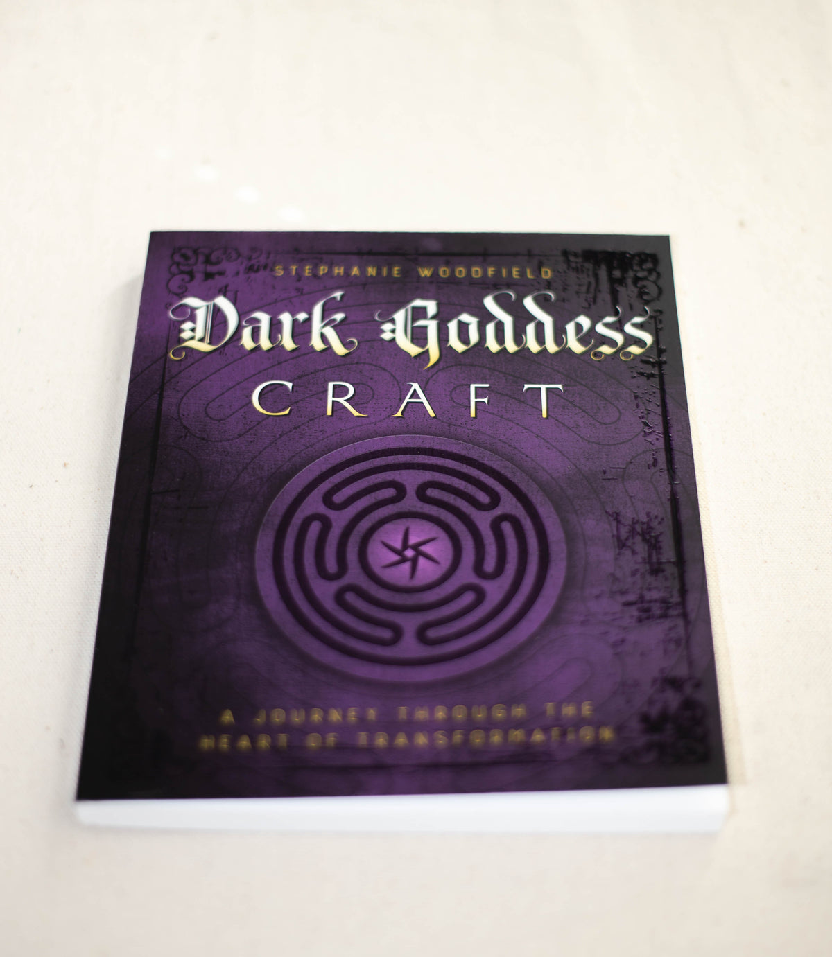 A copy of Dark Goddess Craft by The Healing Hedge Witch rests on a light surface, its dark purple cover featuring a circular labyrinth and gold text. The subtitle reads “A Journey Through the Heart of Transformation.”.