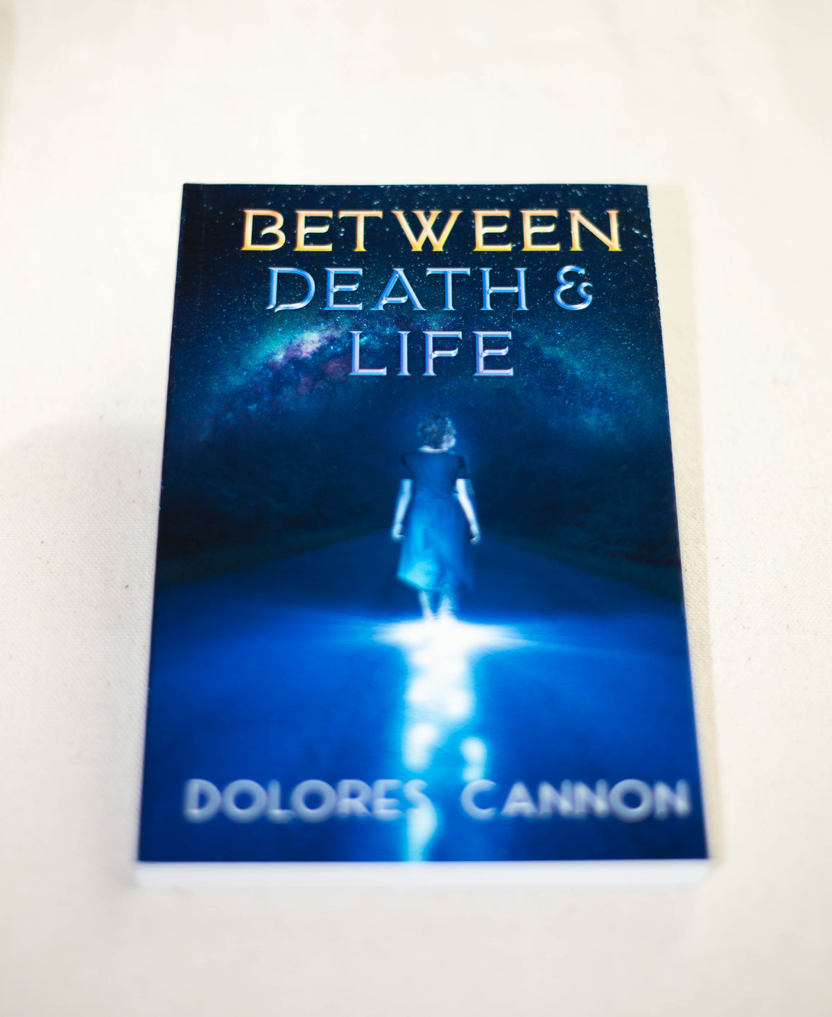 The image shows the book "Between Death and Life" by The Healing Hedge Witch, featuring a person in blue walking on a glowing path beneath a starry sky, with the title and brand name in bold letters.