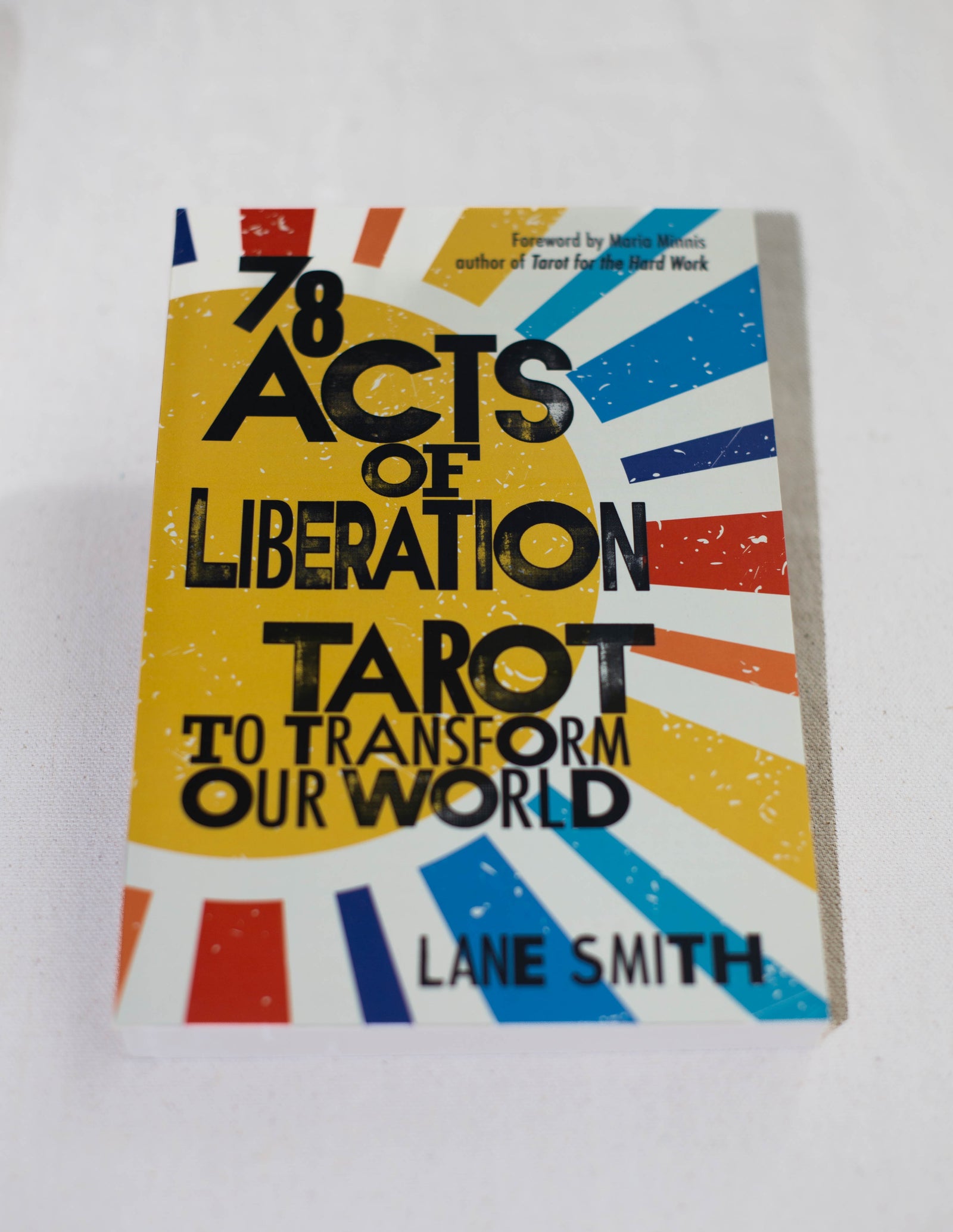 78 Acts of Liberation by The Healing Hedge Witch rests on a white surface, its vibrant cover displaying radiating stripes in red, blue, orange, and white.