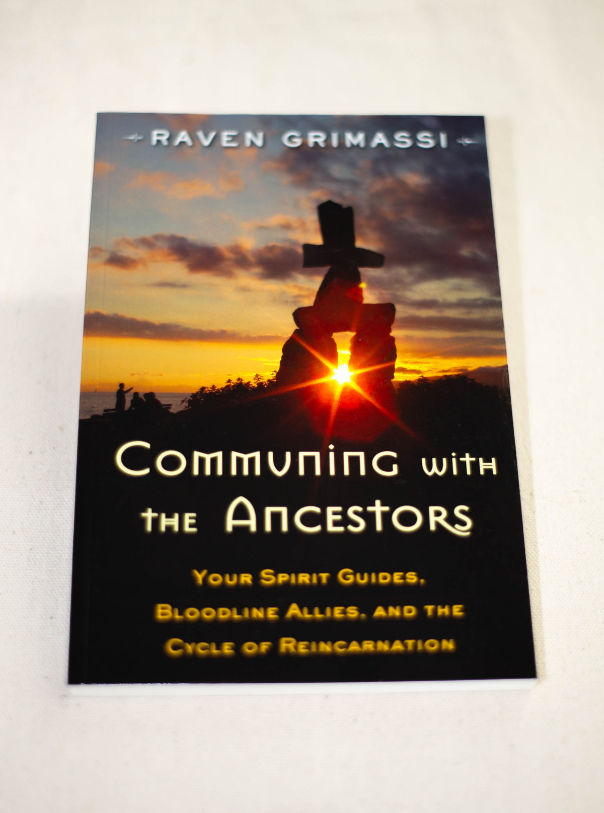Communing with the Ancestors by The Healing Hedge Witch is displayed on a white surface, its cover showing a sunset silhouette of a cross and rocks with sunlight streaming through an opening.
