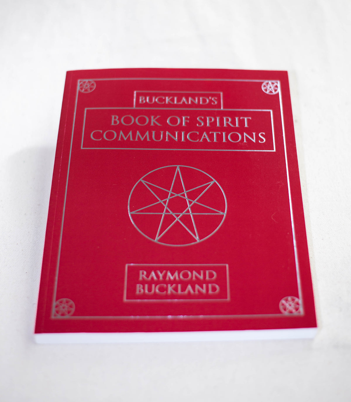 A red book titled "Buckland's Book of Spirit Communications" by The Healing Hedge Witch, featuring a silver pentagram and decorative borders on the cover, is shown lying on a white surface.