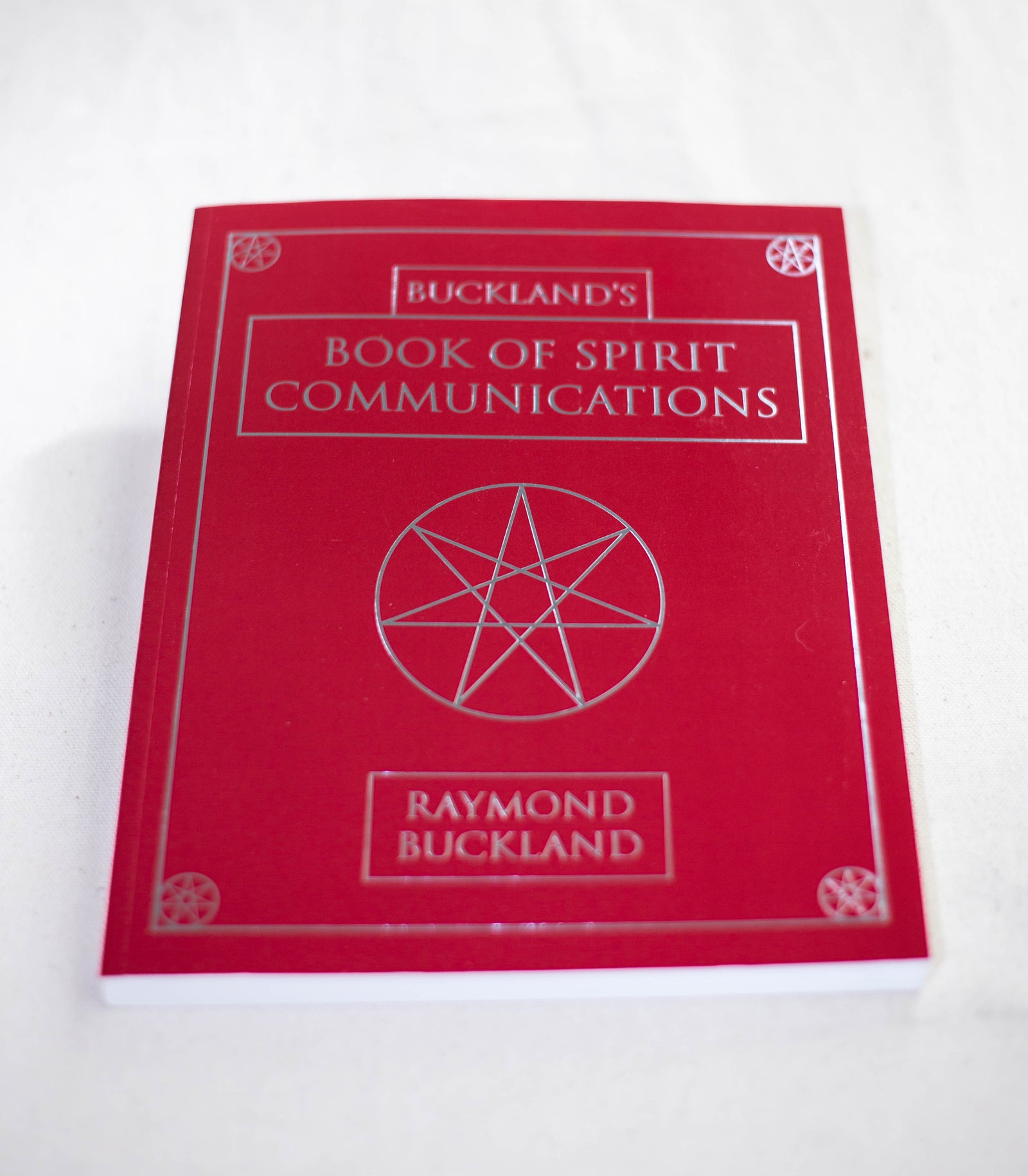 A red book titled "Buckland's Book of Spirit Communications" by The Healing Hedge Witch, featuring a silver pentagram and decorative borders on the cover, is shown lying on a white surface.