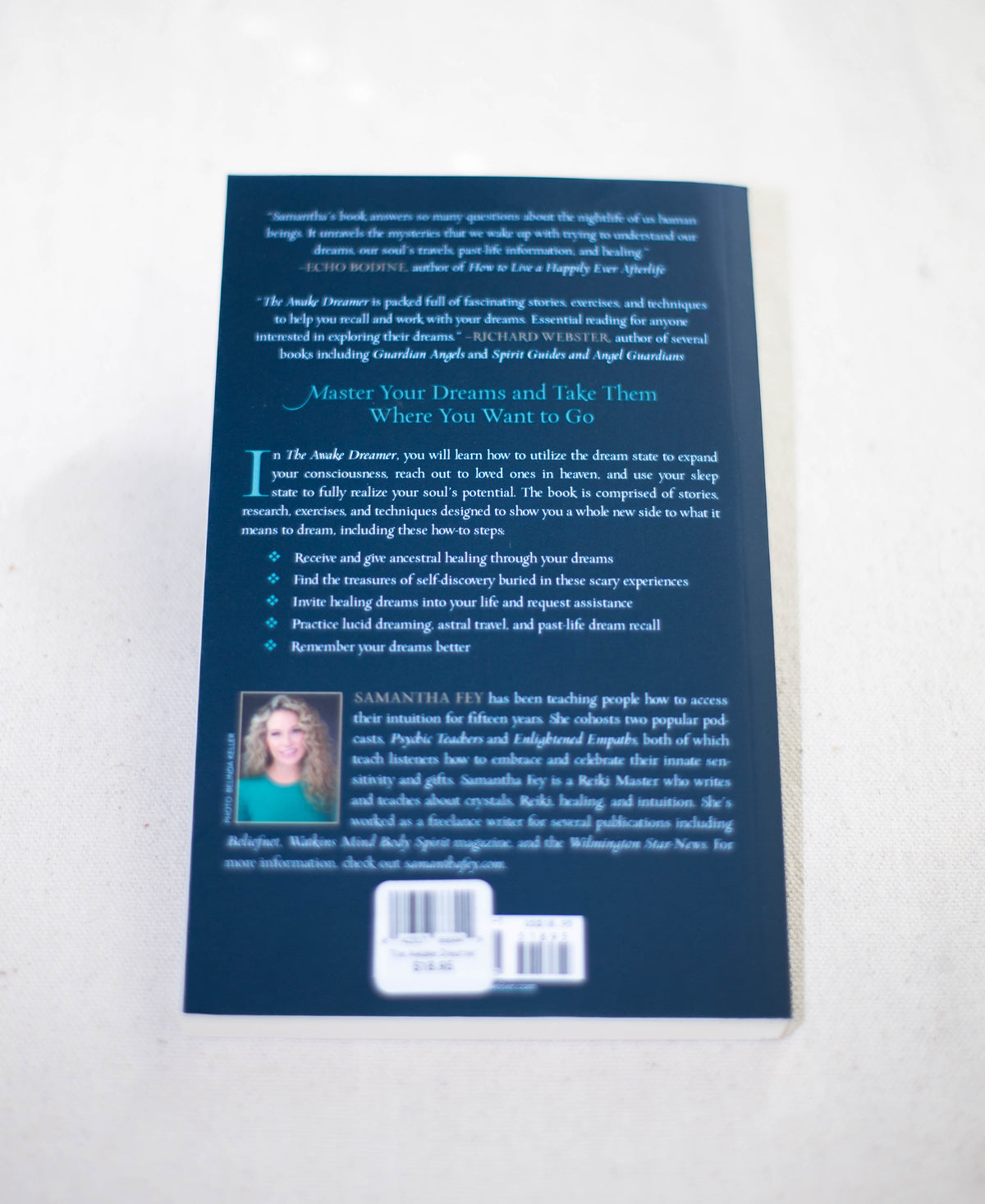 A copy of "The Awake Dreamer" by The Healing Hedge Witch lies face down on a light surface, displaying its dark blue back cover with text, a small woman’s photo in the bottom left corner, and two barcodes near the bottom.