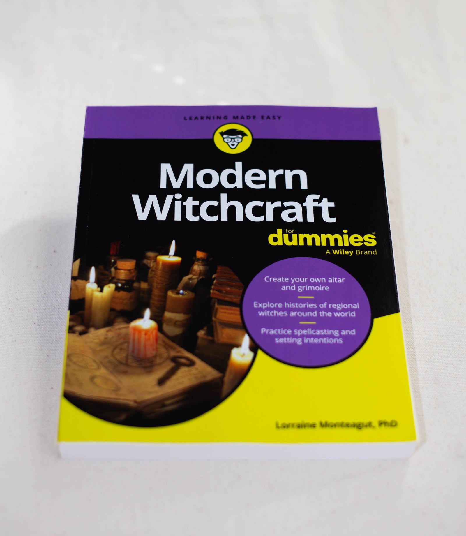 Modern Witchcraft For Dummies by The Healing Hedge Witch rests on a white surface. Its cover displays candles, a crystal ball, spell books, and mystical items, with text about spellcasting, altar making, and the guide’s history.