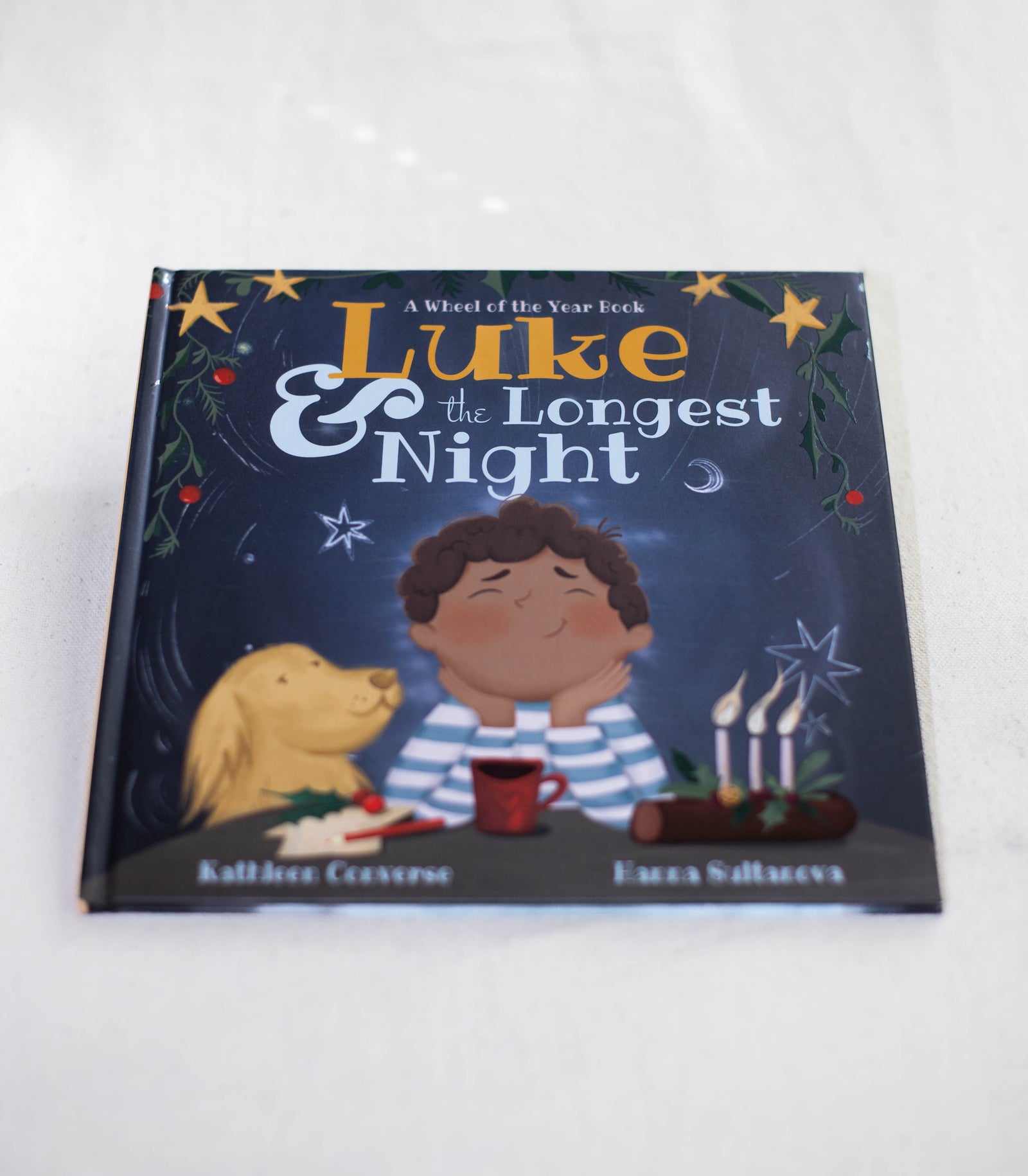 The cover of Luke & the Longest Night by The Healing Hedge Witch features a boy with brown curly hair, a yellow dog, candles, and festive decorations set against a dark blue background.