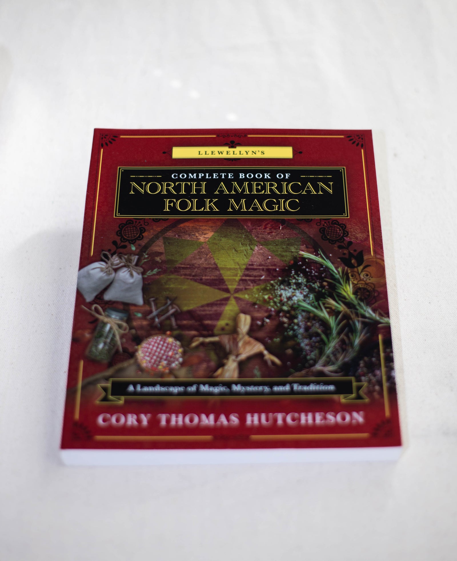 Llewellyn's Complete Book of North American Folk Magic by The Healing Hedge Witch rests on a white surface, its cover decorated with herbs, bottles, and folk items that represent modern folk traditions.