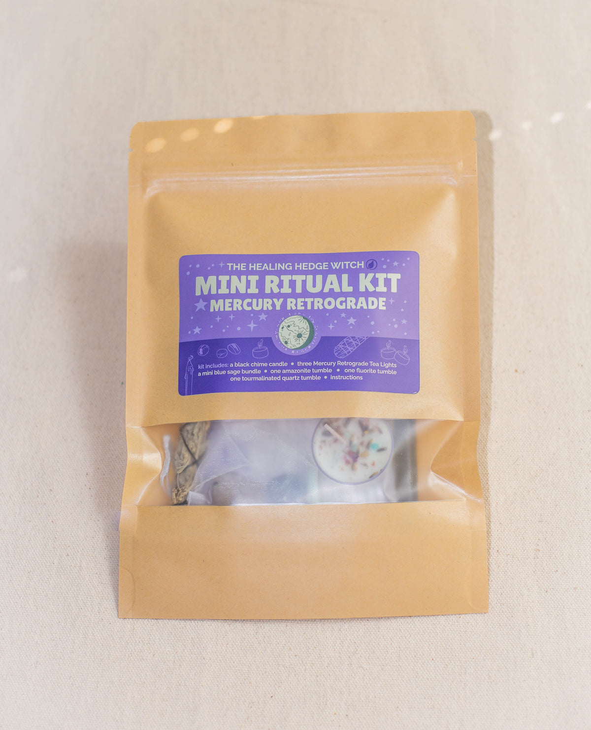A brown resealable pouch labeled Mercury Retrograde Mini Ritual Kit by The Healing Hedge Witch, featuring a purple sticker, contains a tealight candle, herbs, and blue sage bundle for energy protection. It's shown on a light surface.