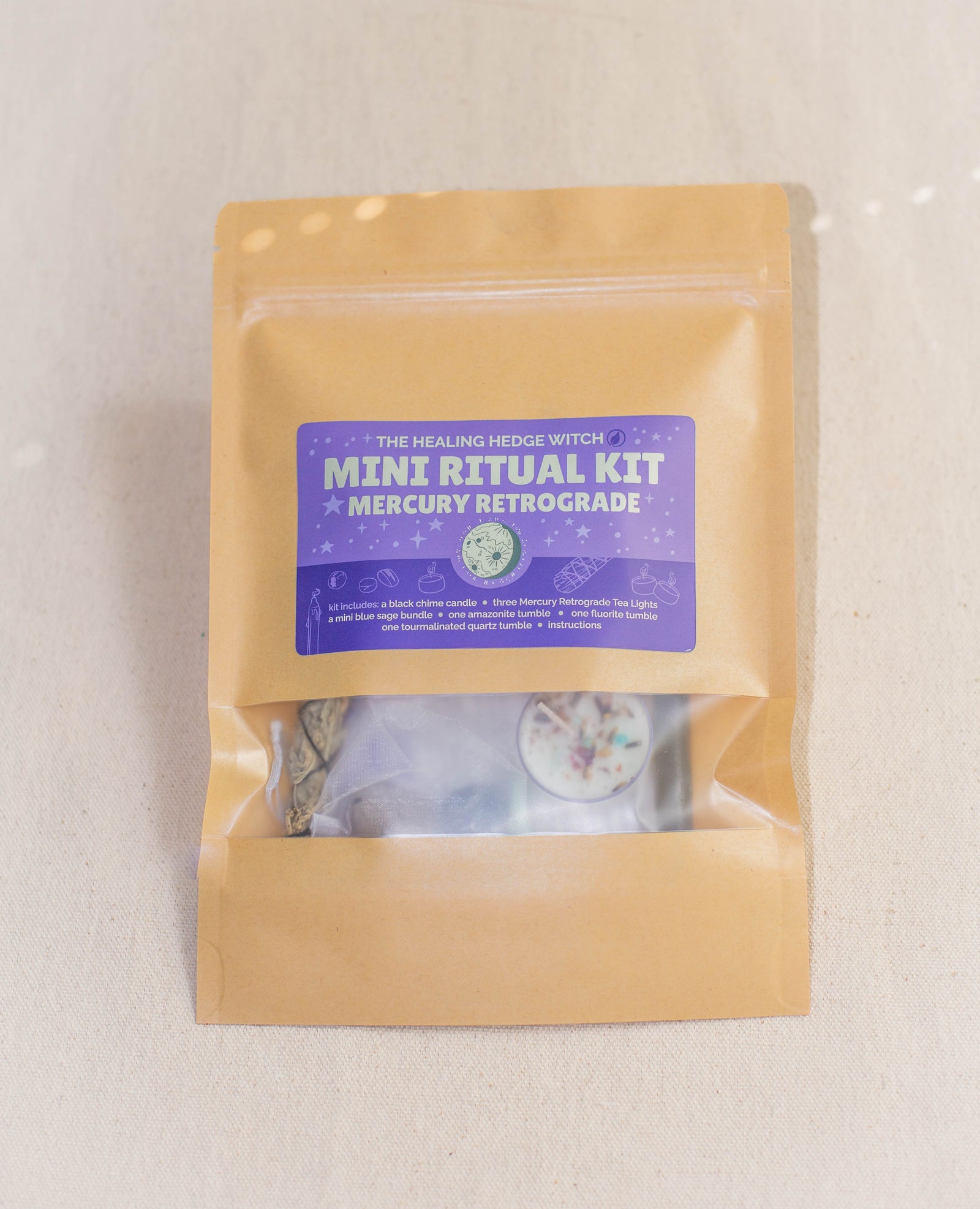 A brown resealable pouch labeled Mercury Retrograde Mini Ritual Kit by The Healing Hedge Witch, featuring a purple sticker, contains a tealight candle, herbs, and blue sage bundle for energy protection. It's shown on a light surface.