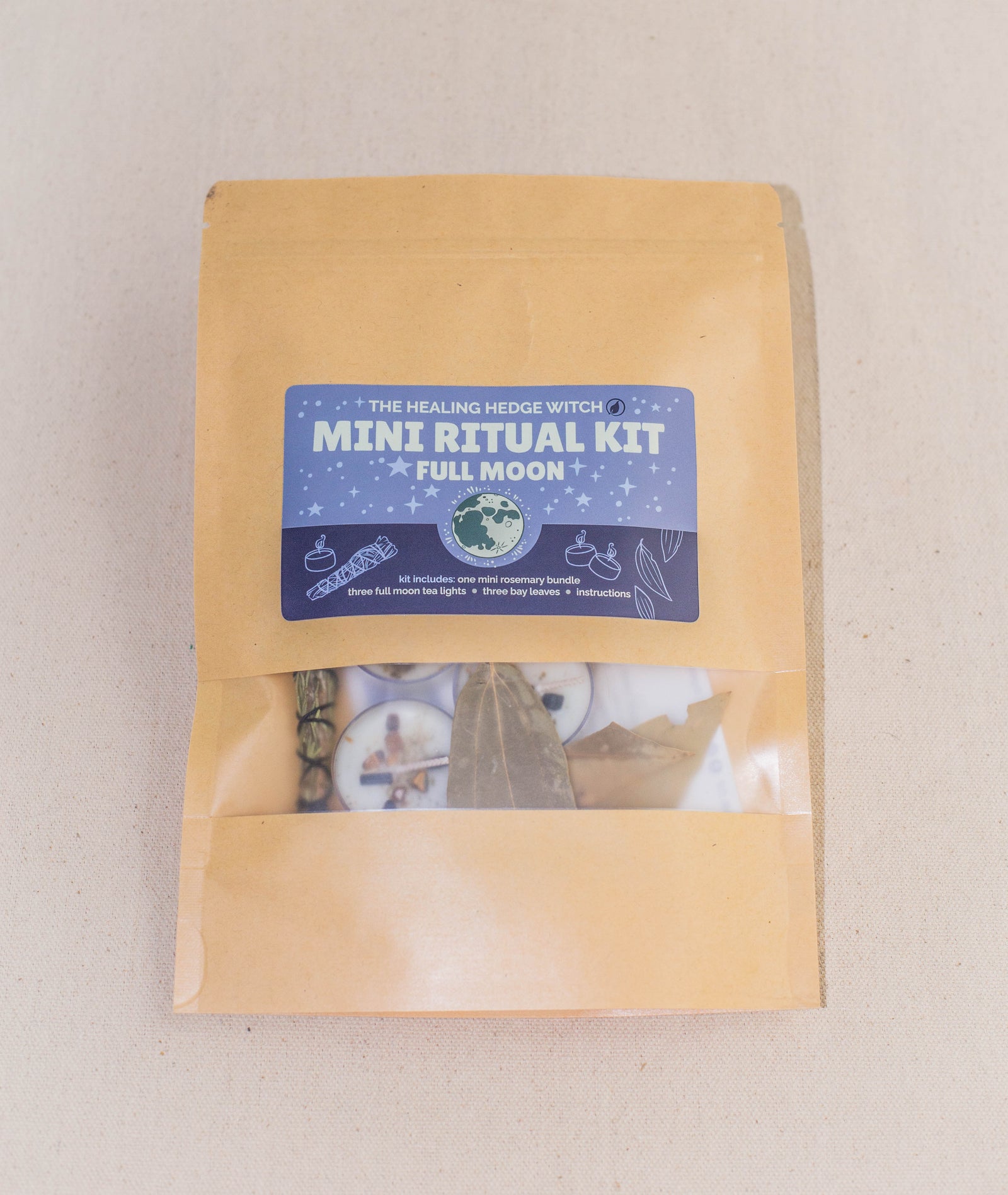 A brown paper package labeled "Full Moon Mini Ritual Kit" by The Healing Hedge Witch features a clear window displaying herbs, a bay leaf, and a rosemary bundle among other ritual items, set on a light beige surface.
