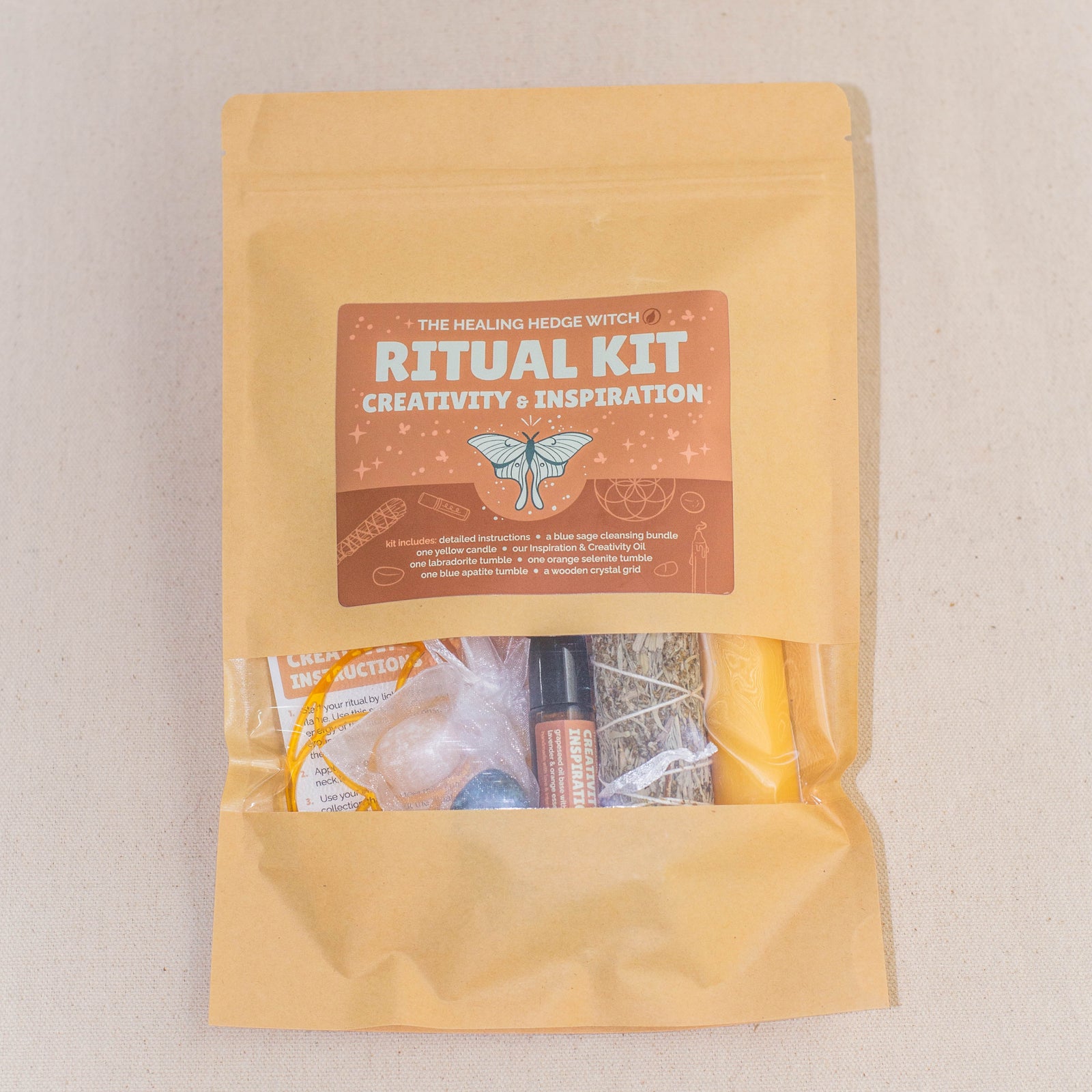 A brown paper pouch labeled Ritual Kit: Creativity & Inspiration contains small items, including crystals, a spray bottle, and herbs, partially visible through a clear window. The pouch is set on a beige fabric surface.