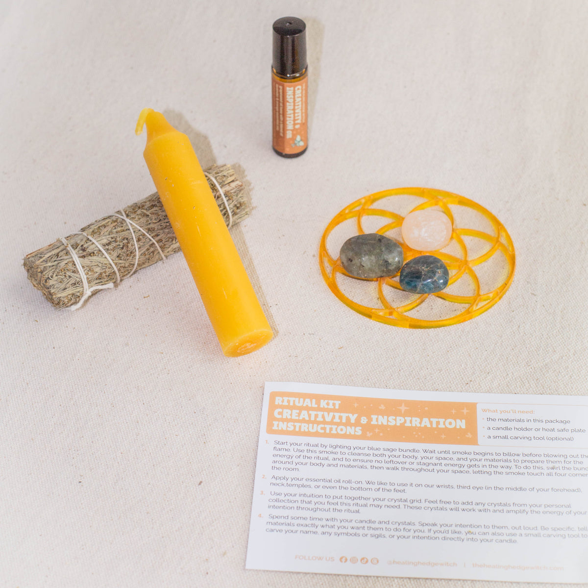 A ritual kit on a light fabric surface, including a yellow candle, sage bundle, essential oil roller, three crystals on a yellow grid, and an instruction card labeled CREATIVITY + INSPIRATION INSTRUCTIONS.