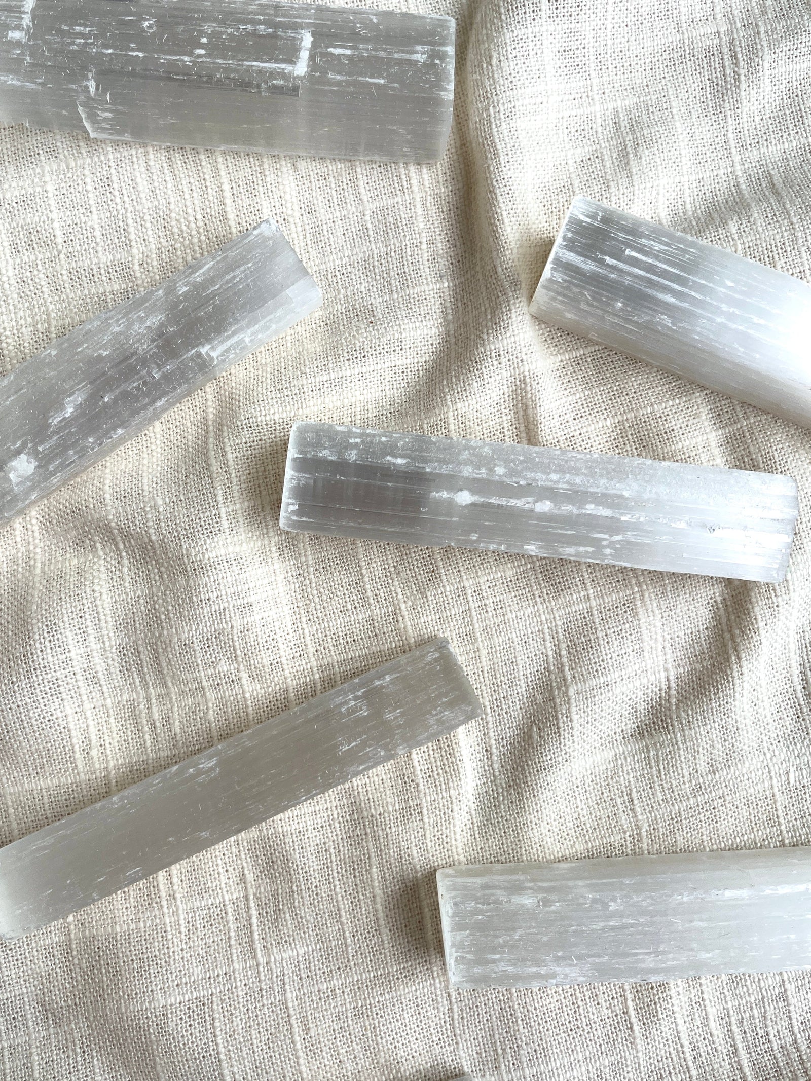 Several translucent, white Selenite Sticks from The Healing Hedge Witch are arranged on a textured cream fabric. These smooth sticks are ideal for crystal protection and cleansing negative energy.