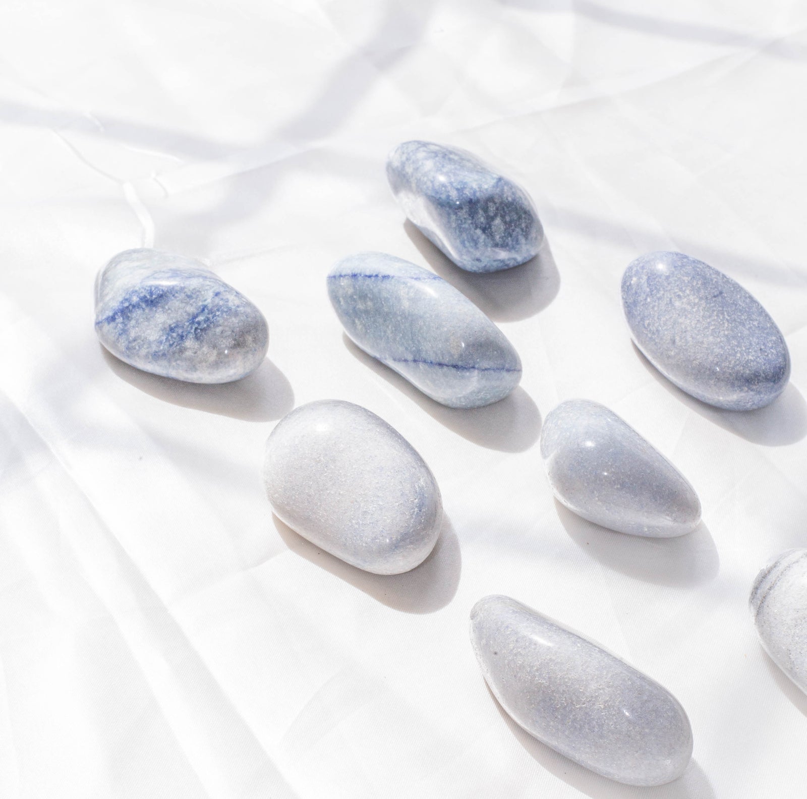 Blue Quartz Palm Stones from The Healing Hedge Witch are displayed on a white fabric background, bathed in soft natural light and shadows for a gentle, serene atmosphere.