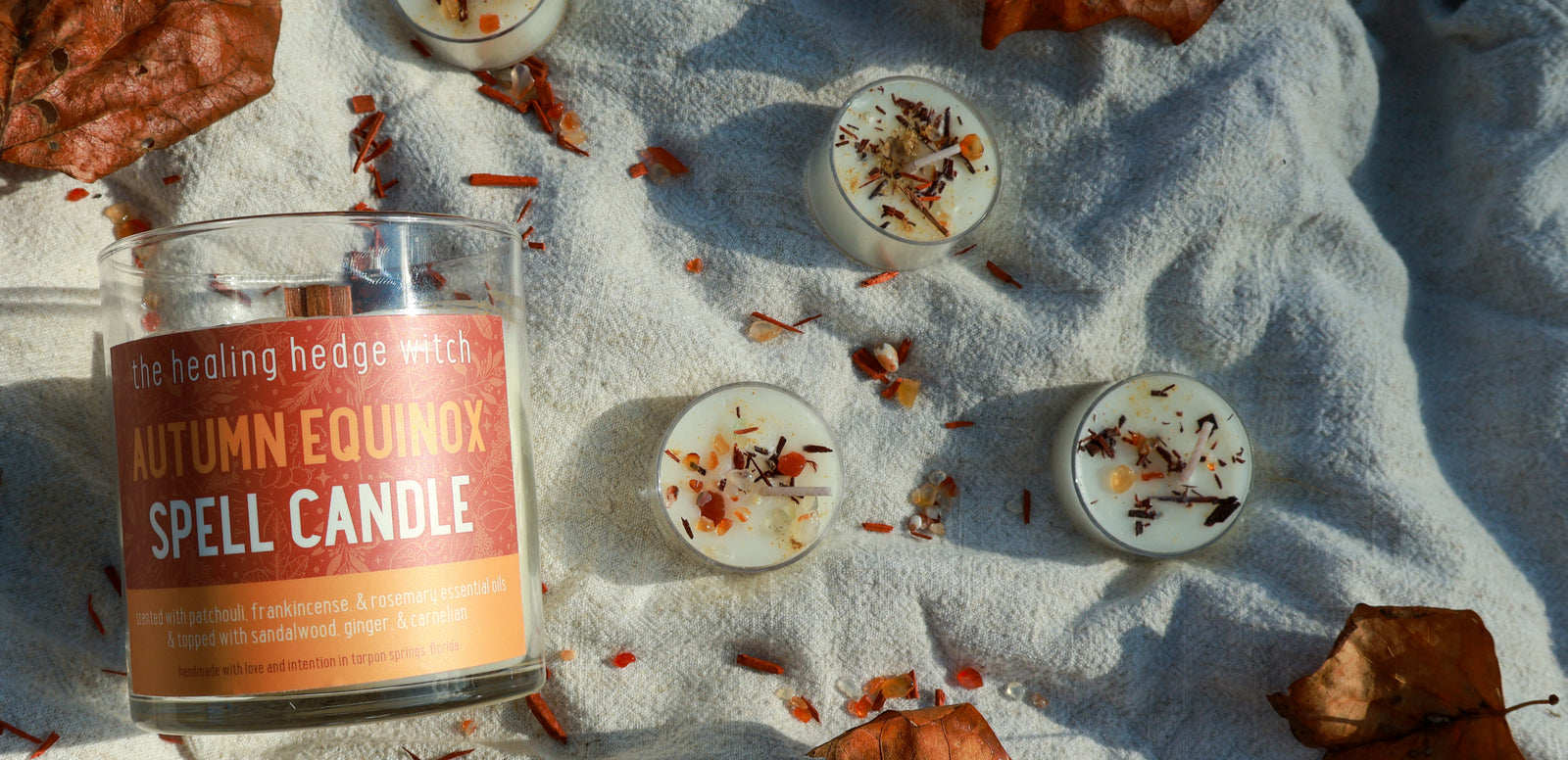 A labeled “Autumn Equinox Spell Candle” sits on a soft, light blanket with several small, decorated tealights and scattered autumn leaves, suggesting a cozy seasonal atmosphere.