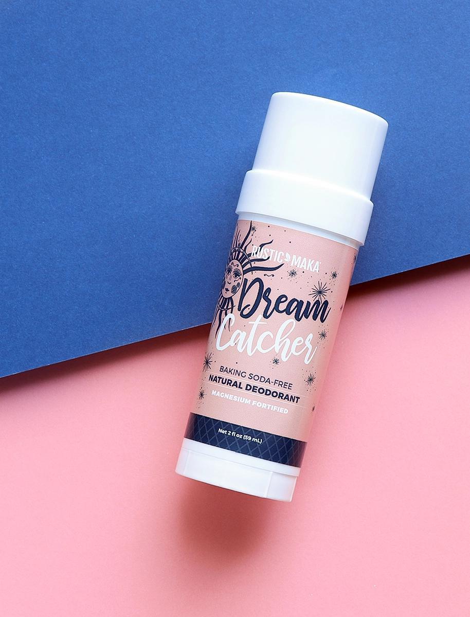 A stick of The Healing Hedge Witch Dream Catcher Natural Deodorant with a white cap sits on a pink and blue background, featuring decorative text and illustrations on the label.