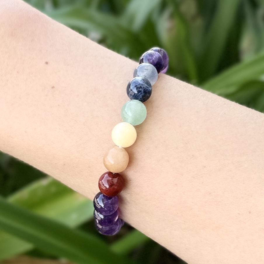 The Healing Hedge Witch Chakra Stone Bracelet, with round multicolored beads, is worn on a wrist against a blurred green backdrop of leaves and plants.