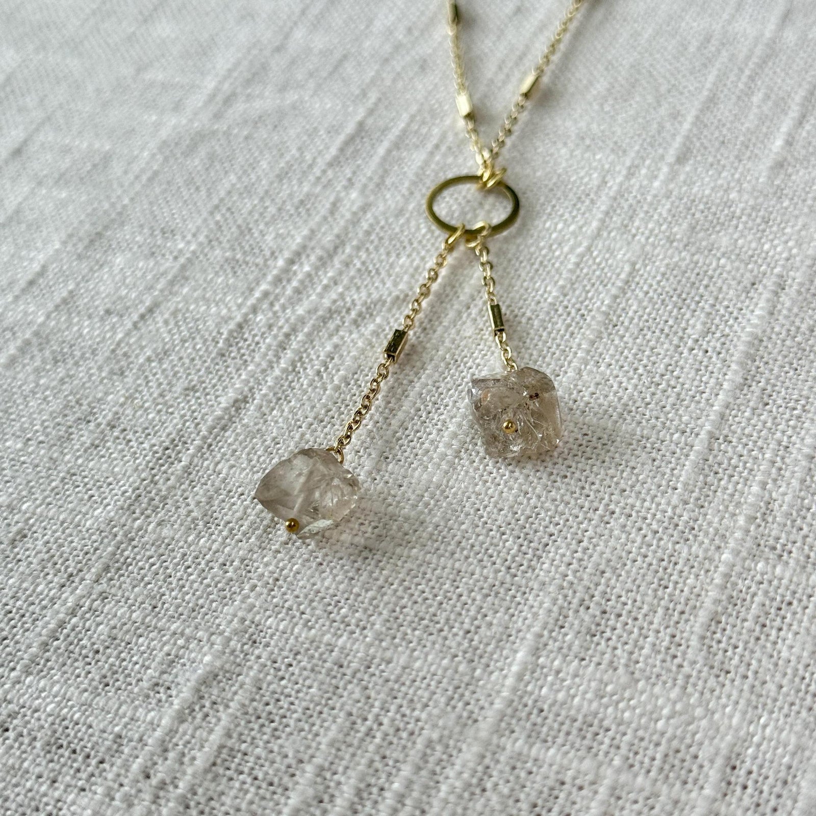 The Healing Hedge Witch's Herkimer Diamond Tassel features two raw, translucent pendants on delicate gold chains, ideal for meditation and crown chakra alignment, shown on a textured white fabric background.