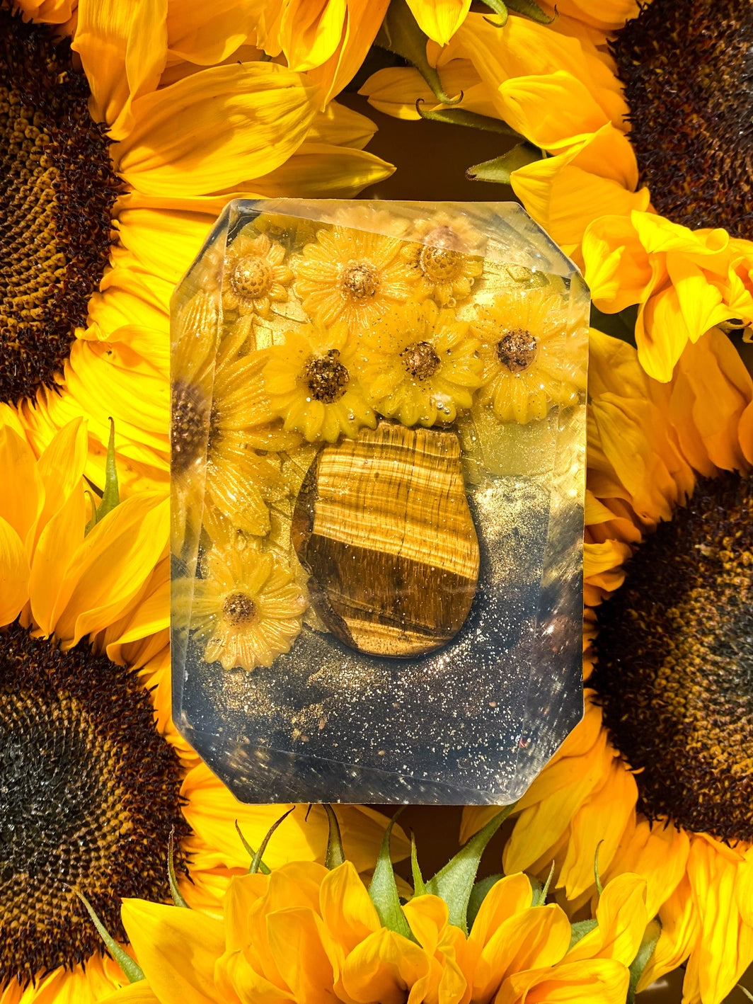 The Healing Hedge Witch's Zodiac Crystal Soap features a rectangular resin block with yellow flowers and a tiger’s eye stone inside, surrounded by vibrant sunflowers with dark centers.