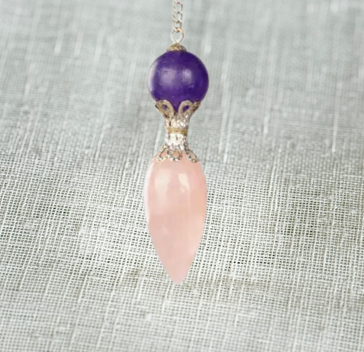 The Healing Hedge Witch’s Rose Quartz with Amethyst Pendulum features a round amethyst bead above a pointed rose quartz, joined by an ornate silver cap—an elegant divination tool displayed on a textured light gray background.