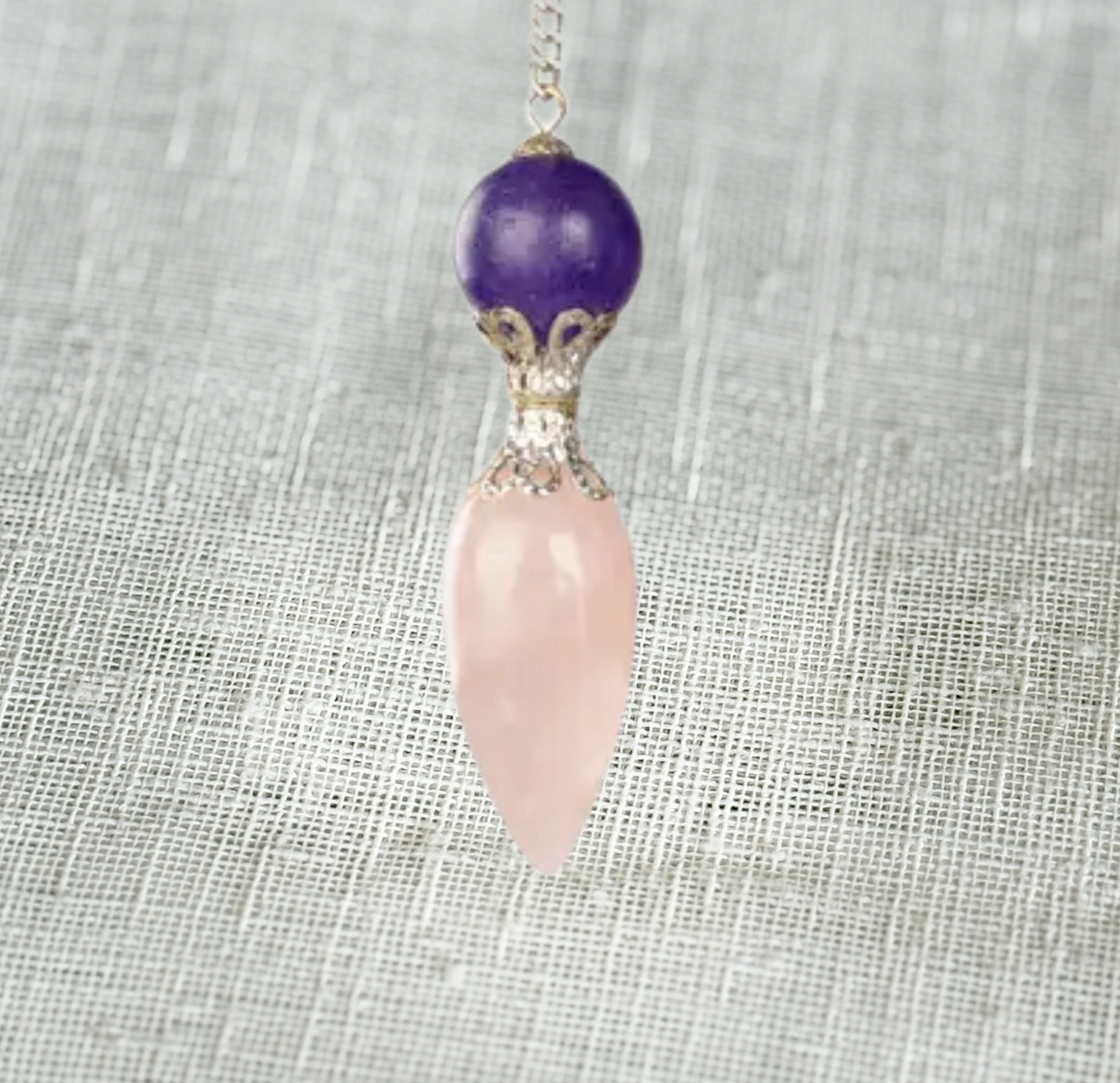 The Healing Hedge Witch’s Rose Quartz with Amethyst Pendulum features a round amethyst bead above a pointed rose quartz, joined by an ornate silver cap—an elegant divination tool displayed on a textured light gray background.