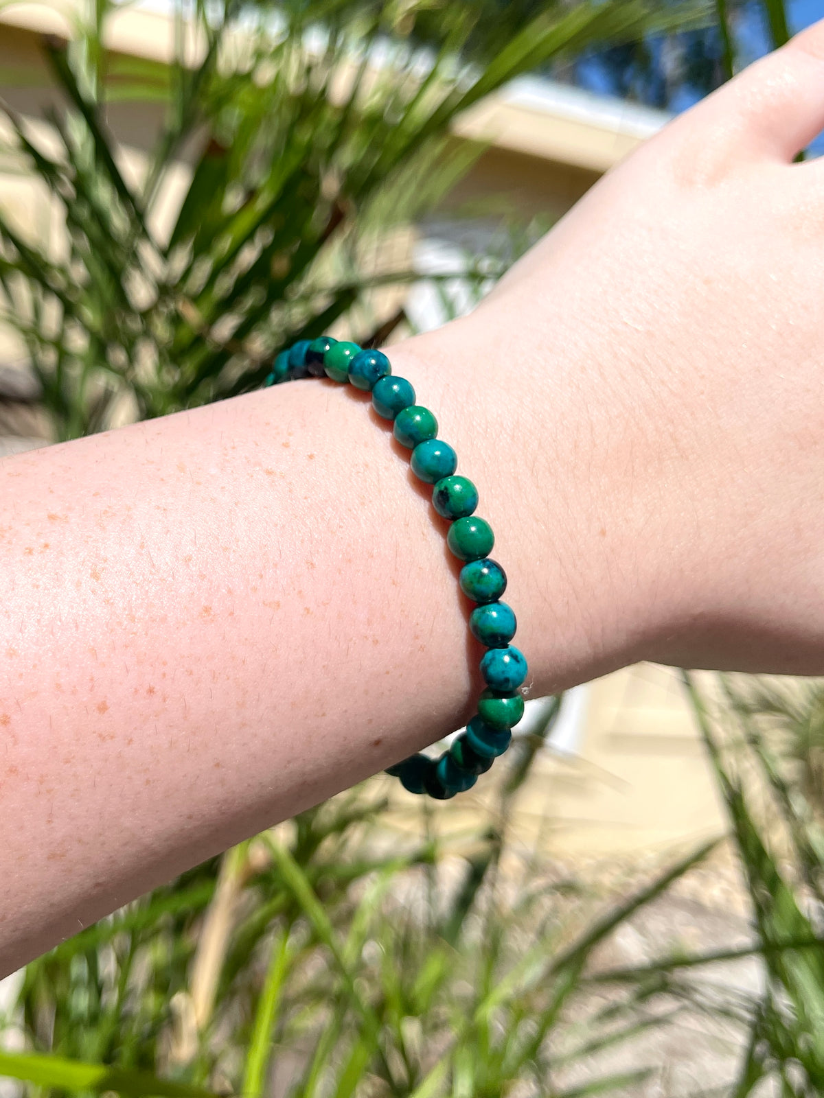 A close-up shows a wrist wearing The Healing Hedge Witch’s Chrysocolla Crystal Bracelet, featuring round greenish-blue beads, with leafy plants and sunlight in the background—ideal for energy healing enthusiasts.