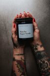 A person with tattooed arms and red nail polish holds a black Maven Made - Faire Matcha Sugar Scrub jar, an antioxidant-rich exfoliant, against a neutral background.