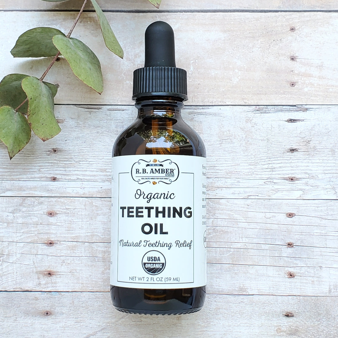 A brown glass dropper bottle labeled R.B. Amber Jewelry - Faire Organic Teething Oil for Safe + Effective Teething Relief sits on a light wood surface with green leaves in the top left corner.